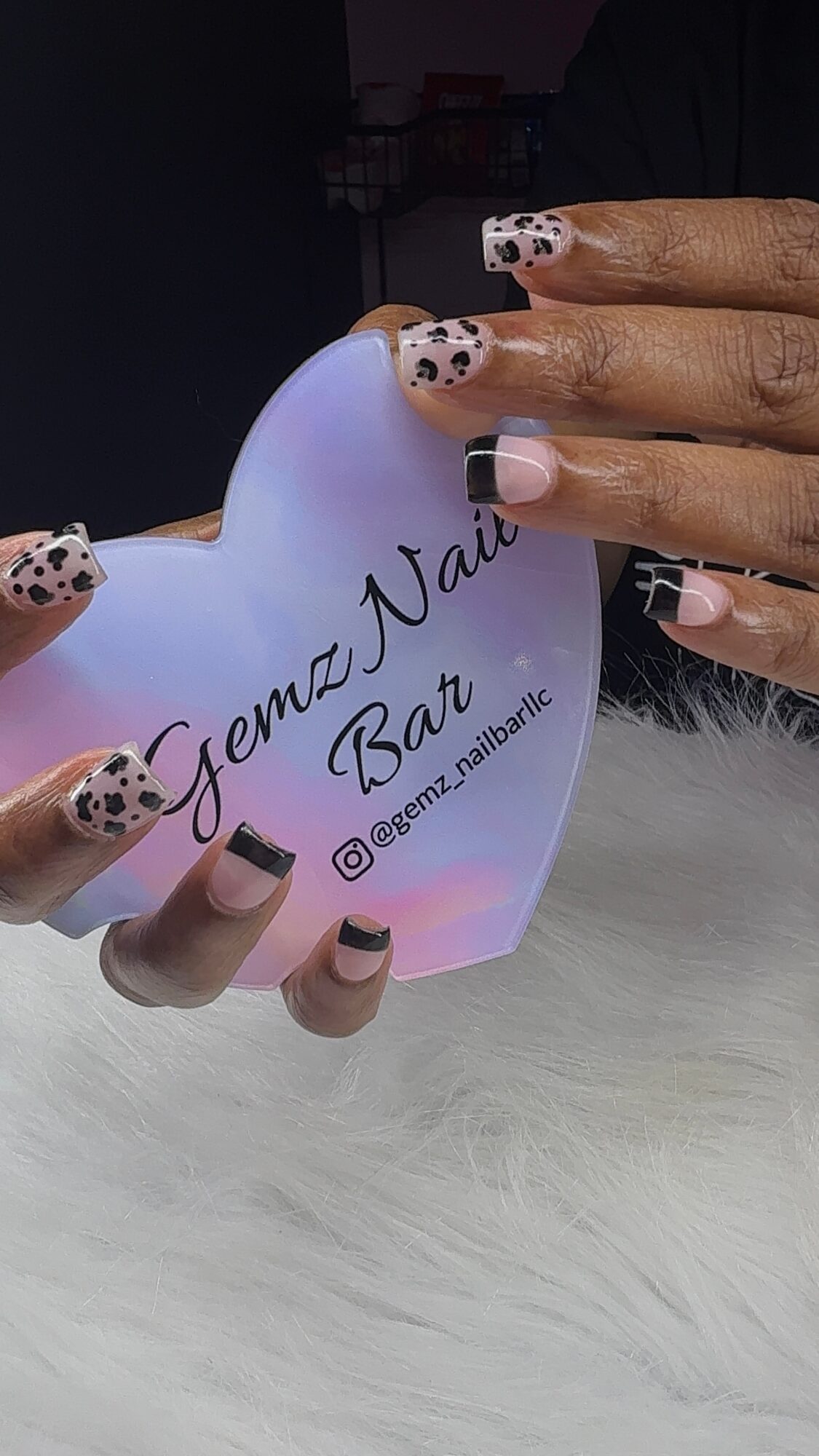 Person holding a heart-shaped card with text, nails painted with black and white spotted design, on a white furry surface.