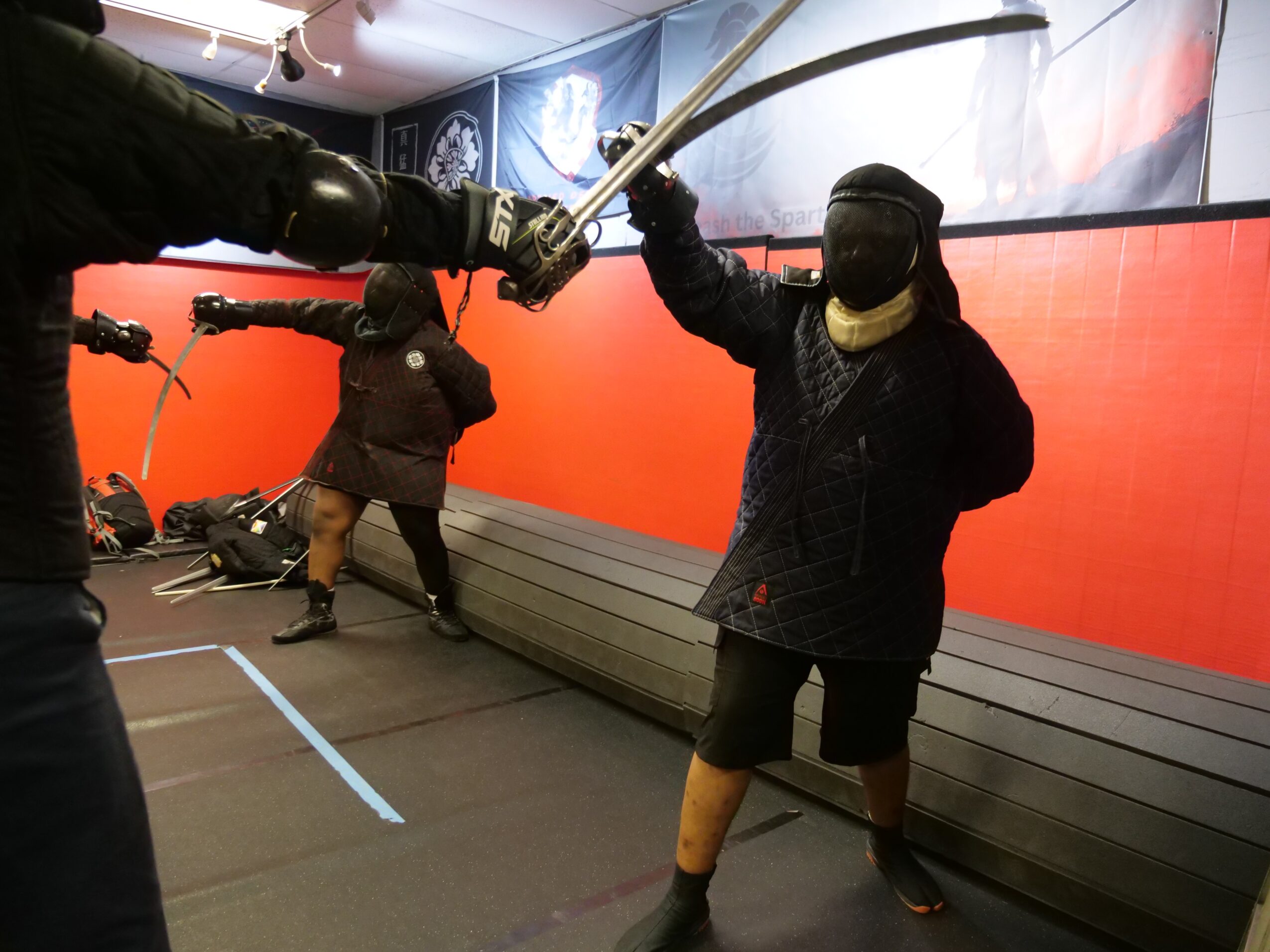 Two people in protective gear hold foam swords during a fencing activity indoors, with a red wall and benches in the background.