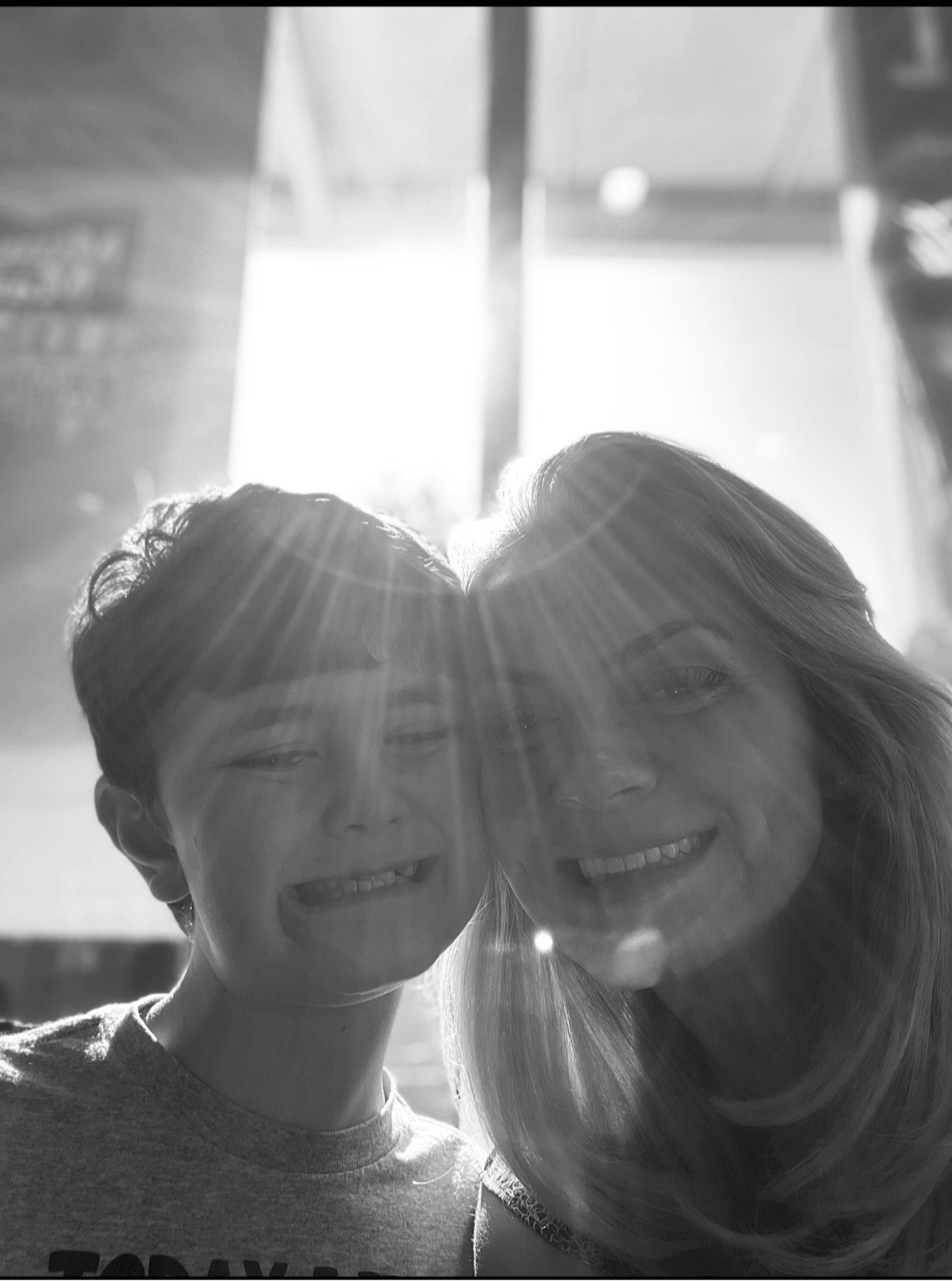 Two people smiling close together with sunlight behind them, creating lens flare and backlighting, in black and white.