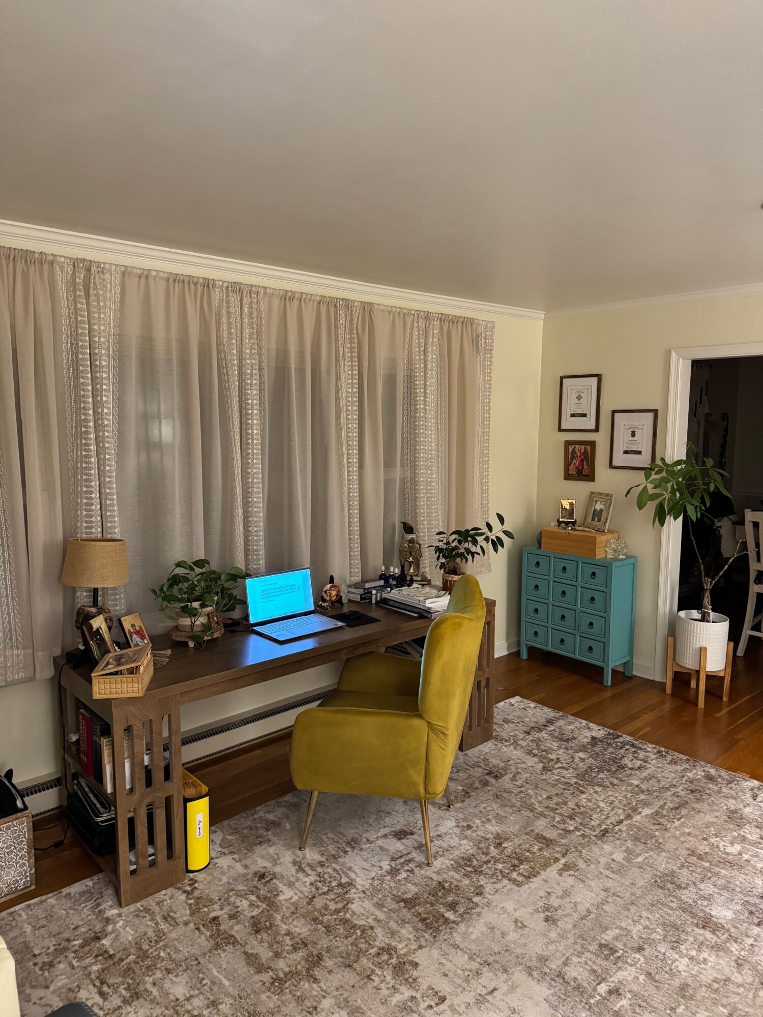 Living room with a desk, yellow chair, blue cabinet, plants, and a window with curtains.