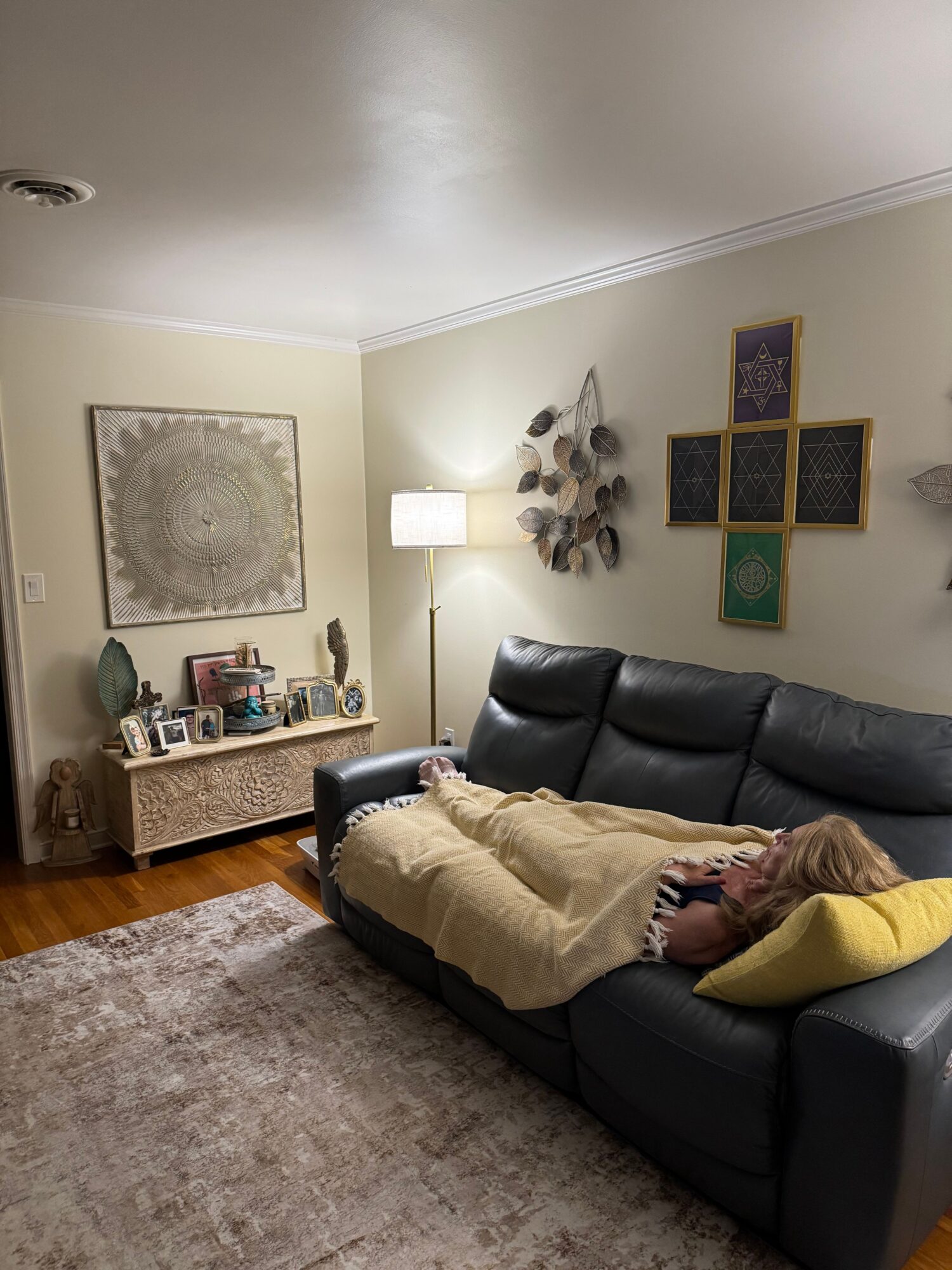 Person lying on a dark sofa with a blanket and yellow pillow in a living room, decorated with wall art and a lamp.