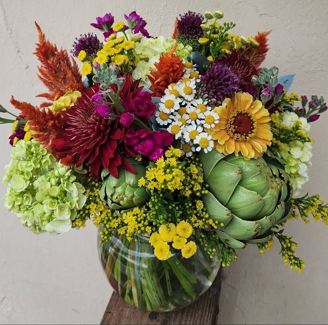 Colorful bouquet of various flowers including daisies, sunflowers, and greenery in a glass vase on a wooden surface.