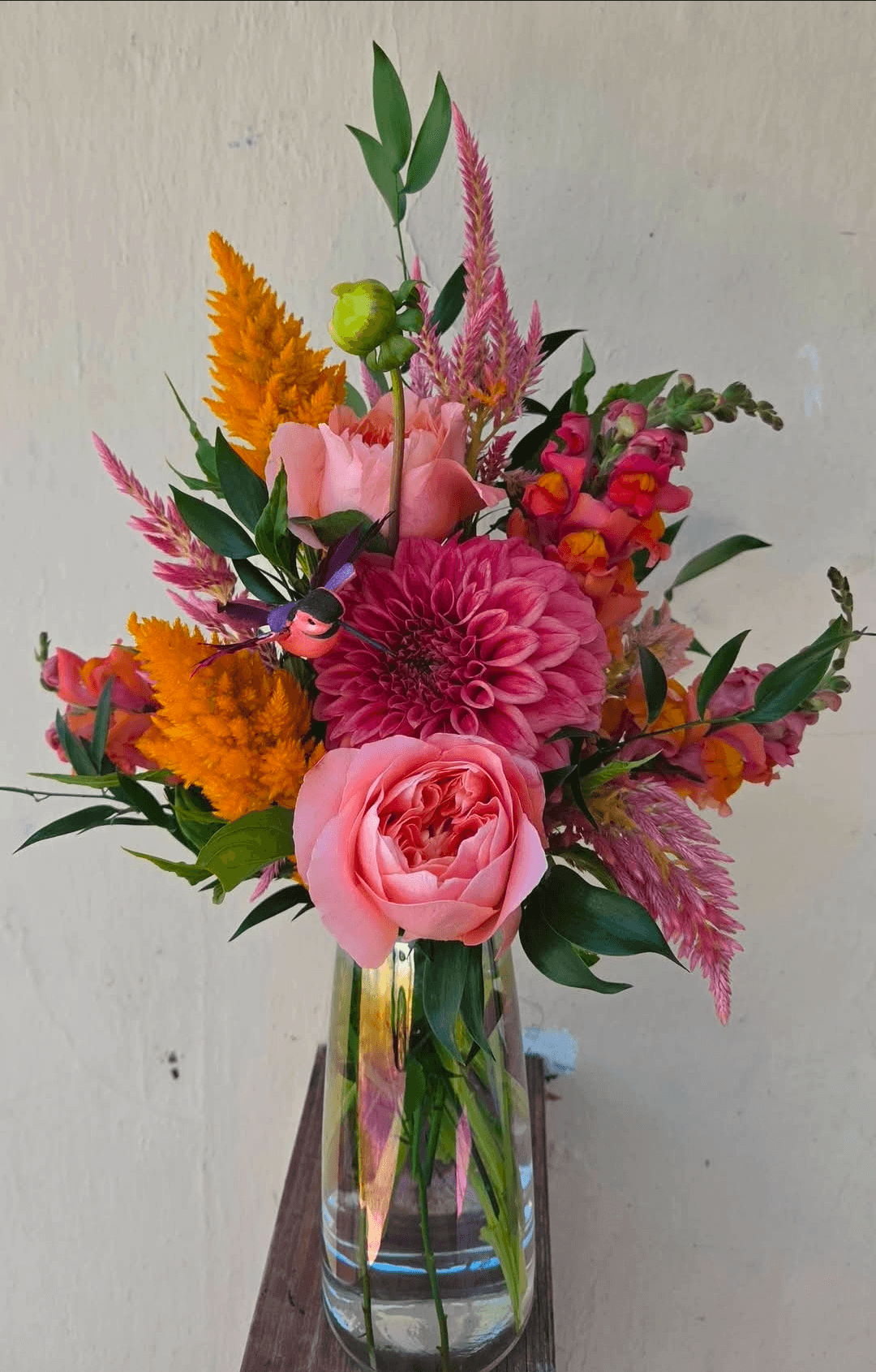 Colorful flower bouquet in a glass vase with pink roses, orange, pink, and purple flowers, and green foliage.