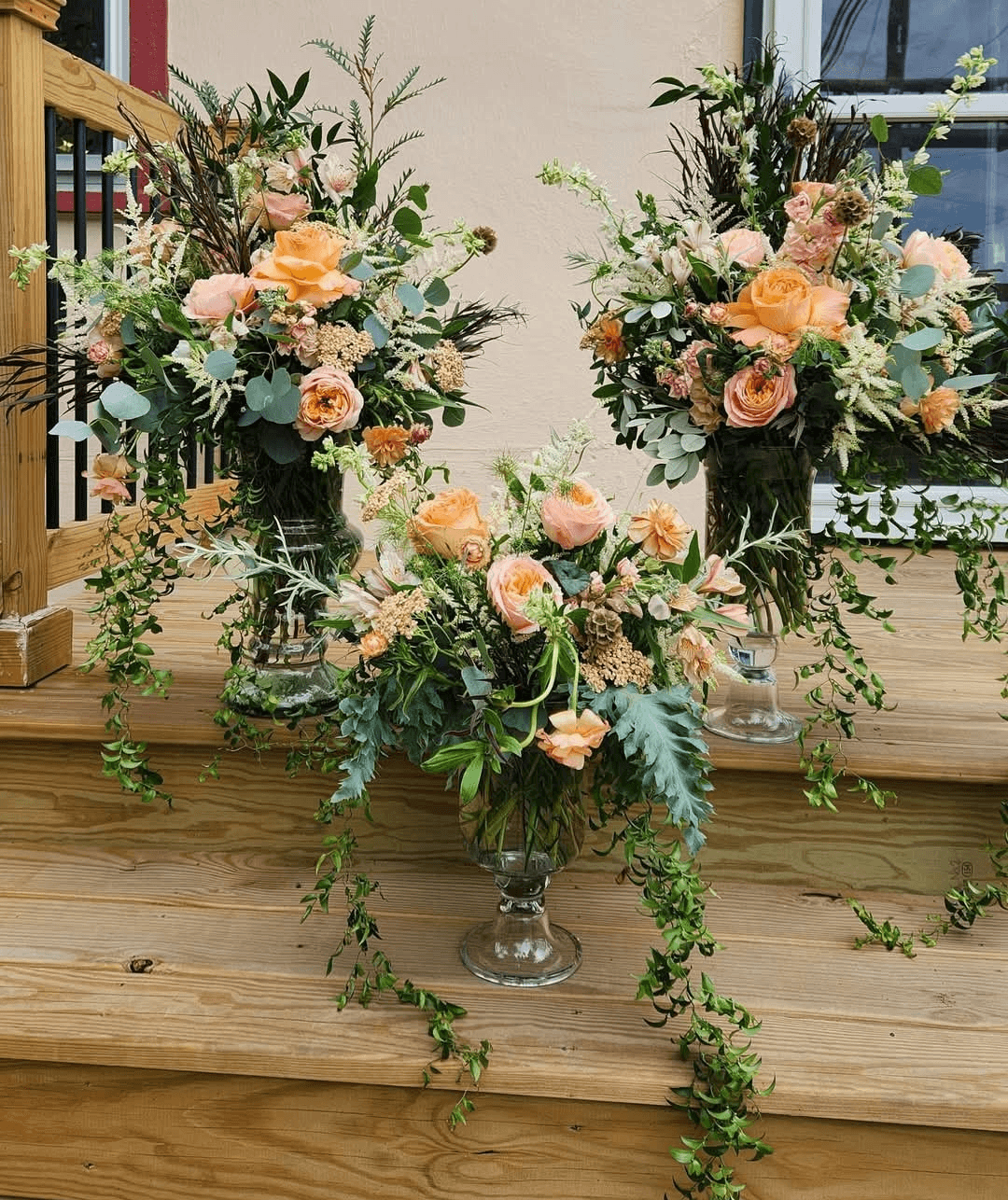 Three floral arrangements with pink, peach, and white flowers and green foliage on wooden steps, outdoors.