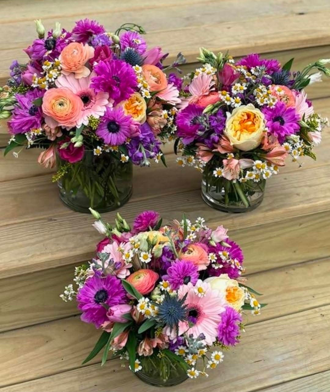 Three colorful flower arrangements in glass vases on a wooden surface, featuring pink, purple, yellow, and white flowers.