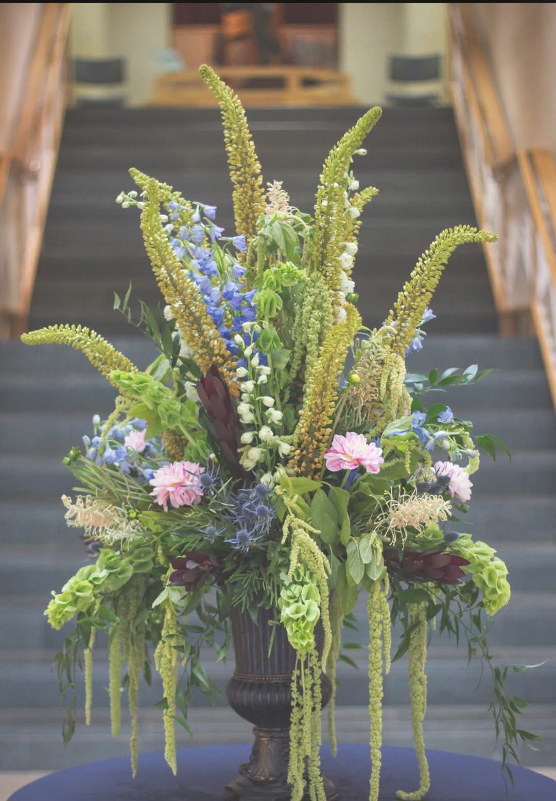 Flower arrangement with tall green and purple flowers in a dark vase on stairs, background blurred.