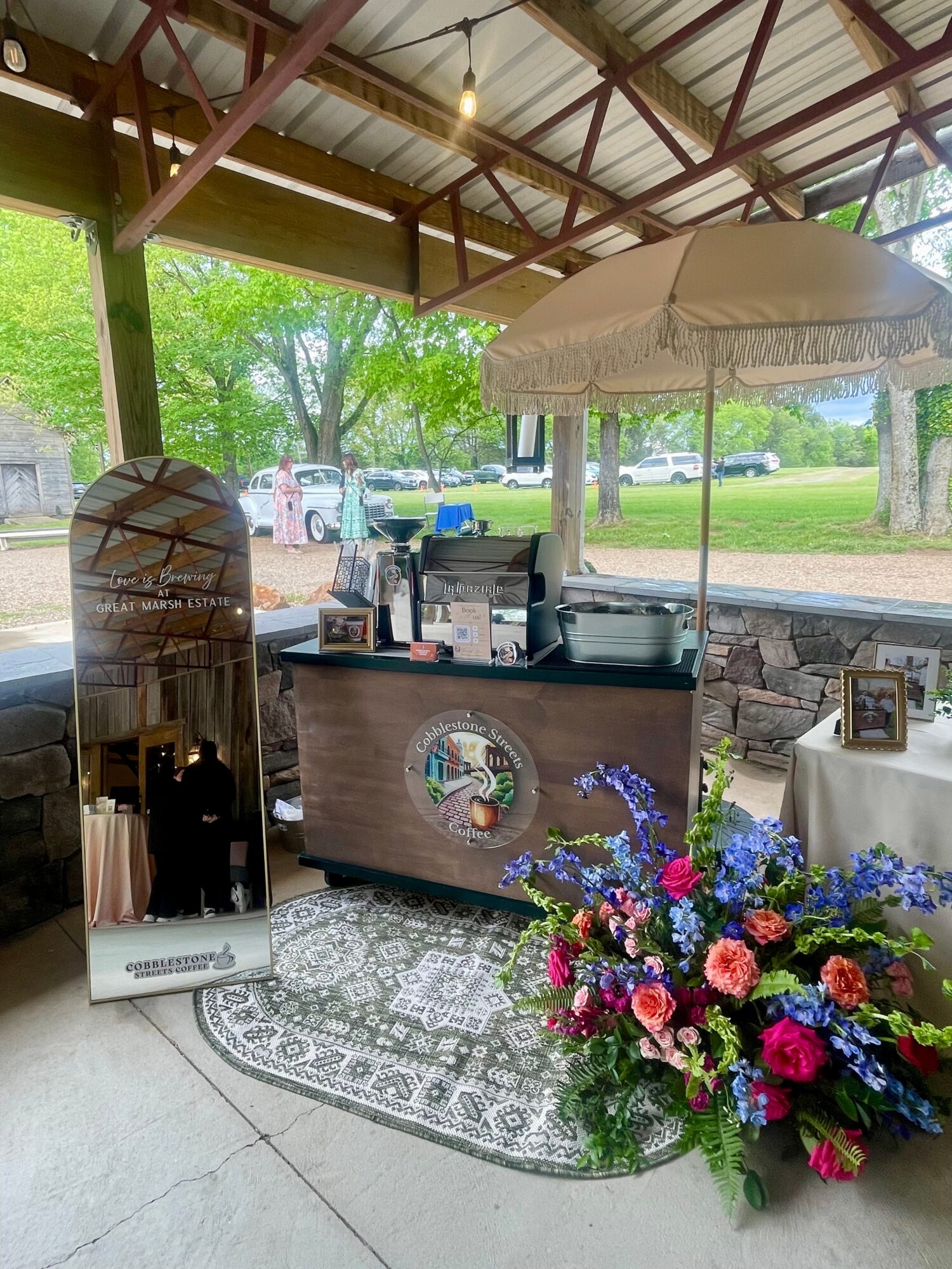 A small outdoor kiosk with a wooden front, decorated with a floral arrangement, under a canopy with a white umbrella, surrounded by greenery.