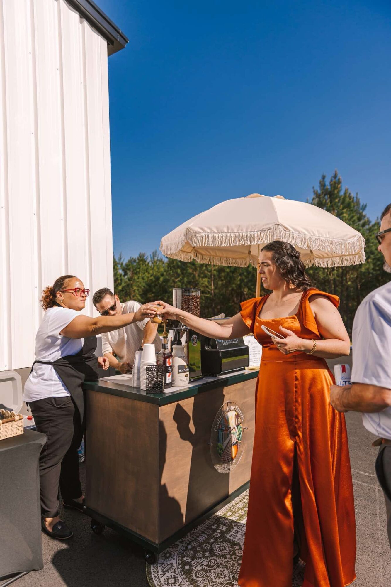 Two women exchanging drinks outdoors under a large umbrella, with a white building and blue sky in the background.