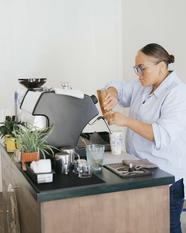 Woman in glasses and striped shirt operating coffee machine in a kitchen, with plants and utensils nearby.
