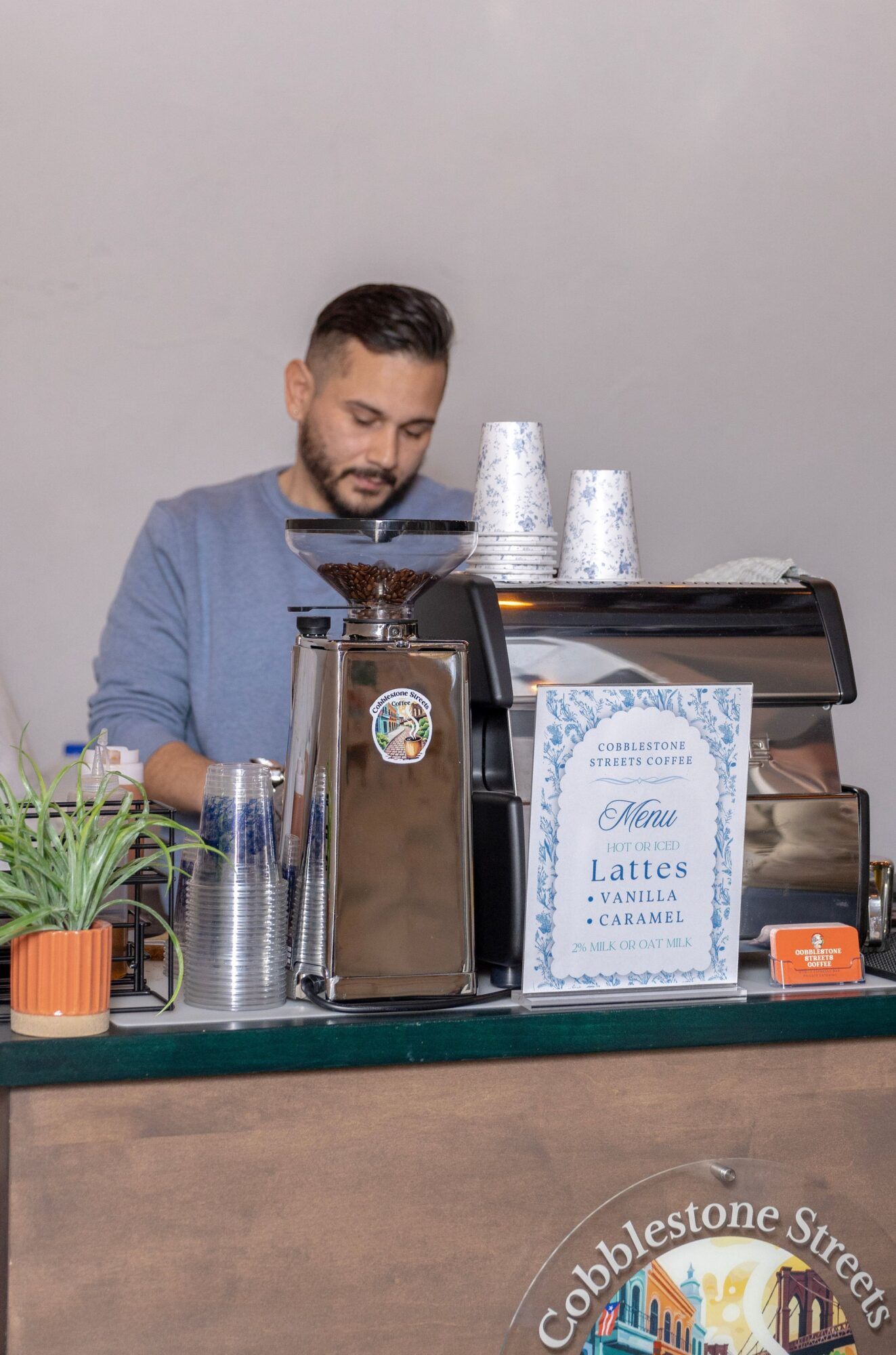 Barista preparing coffee behind a counter with a coffee machine, cups, and a sign listing latte options, in a neutral setting.