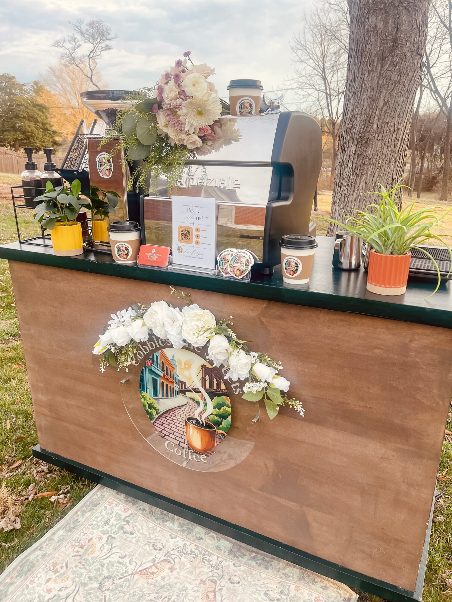 Outdoor wooden counter with flowers, coffee cups, and a coffee machine, surrounded by trees and a grassy area.
