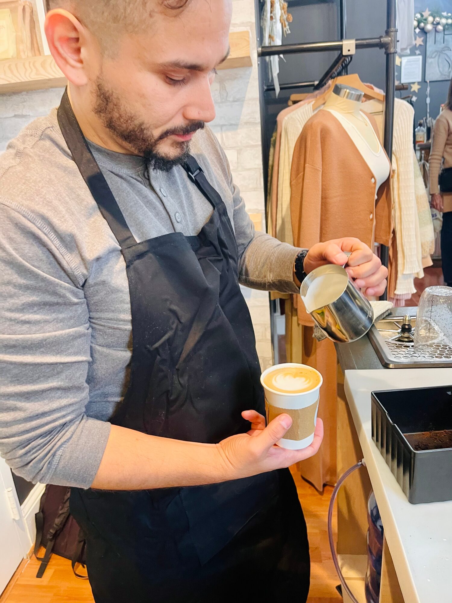 Barista pours steamed milk into a coffee cup in a cafe, with clothing racks in the background.