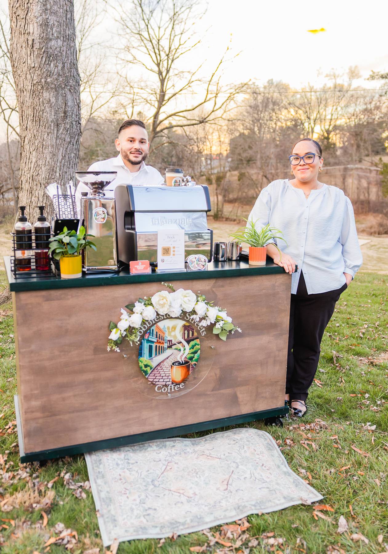 Two people stand behind a coffee stand outdoors with trees and a fence in the background. The stand has a colorful logo and flowers.