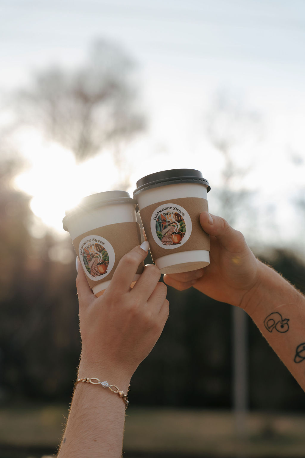 Two hands holding coffee cups outdoors with trees and sky in the background.