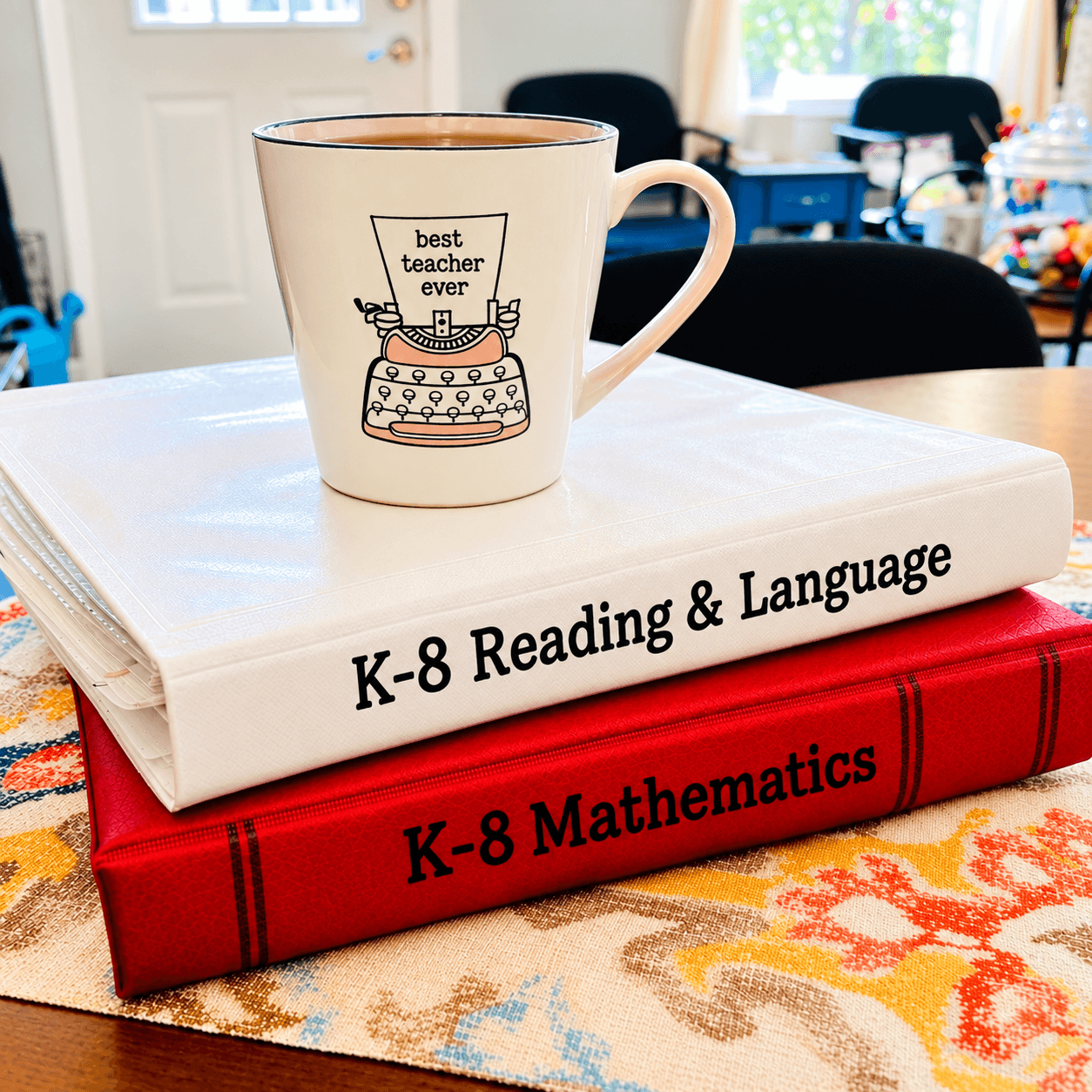 A white mug with a cake illustration and text, placed on two stacked books about K-8 reading and mathematics, on a table.