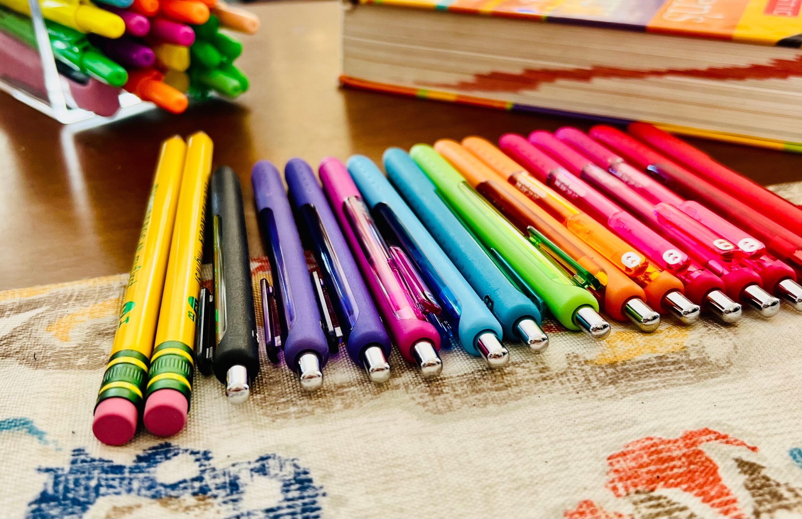 Colorful markers arranged in a row on a table with books and other markers in the background.