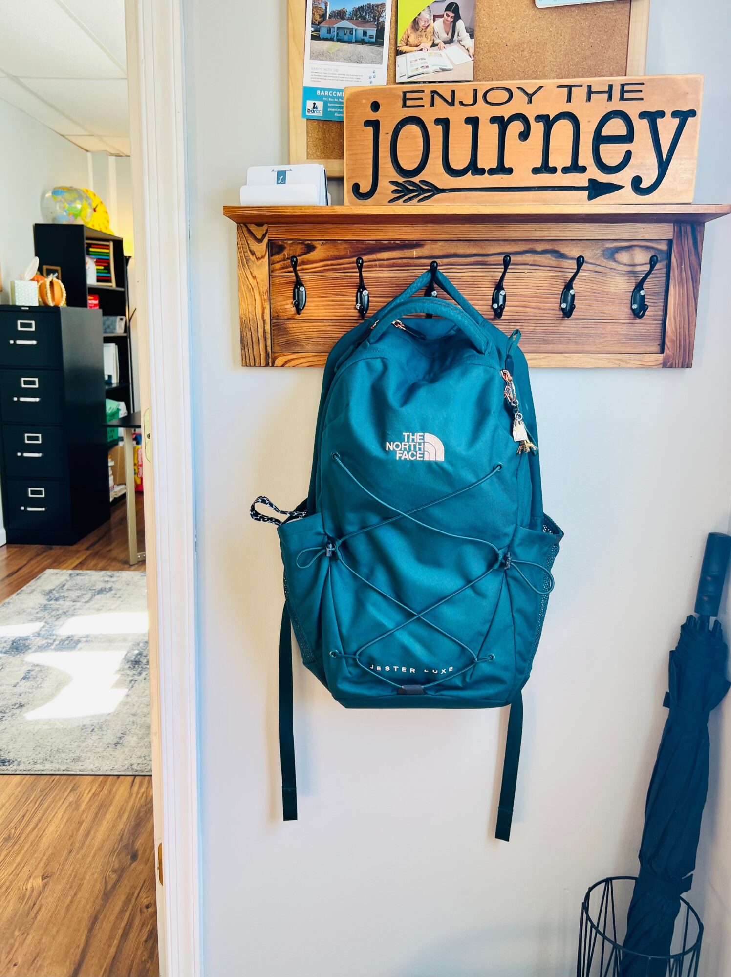 Blue backpack hanging on a wooden hook rack with a sign reading 'ENJOY THE journey' above it, in a room with a door and furniture.
