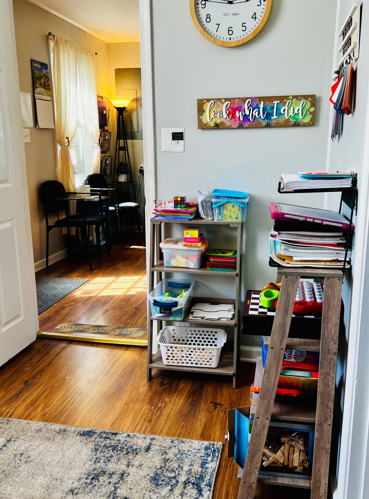 View of a room with a doorway leading to a dining area, wall clock, colorful sign, and shelves with books and items.