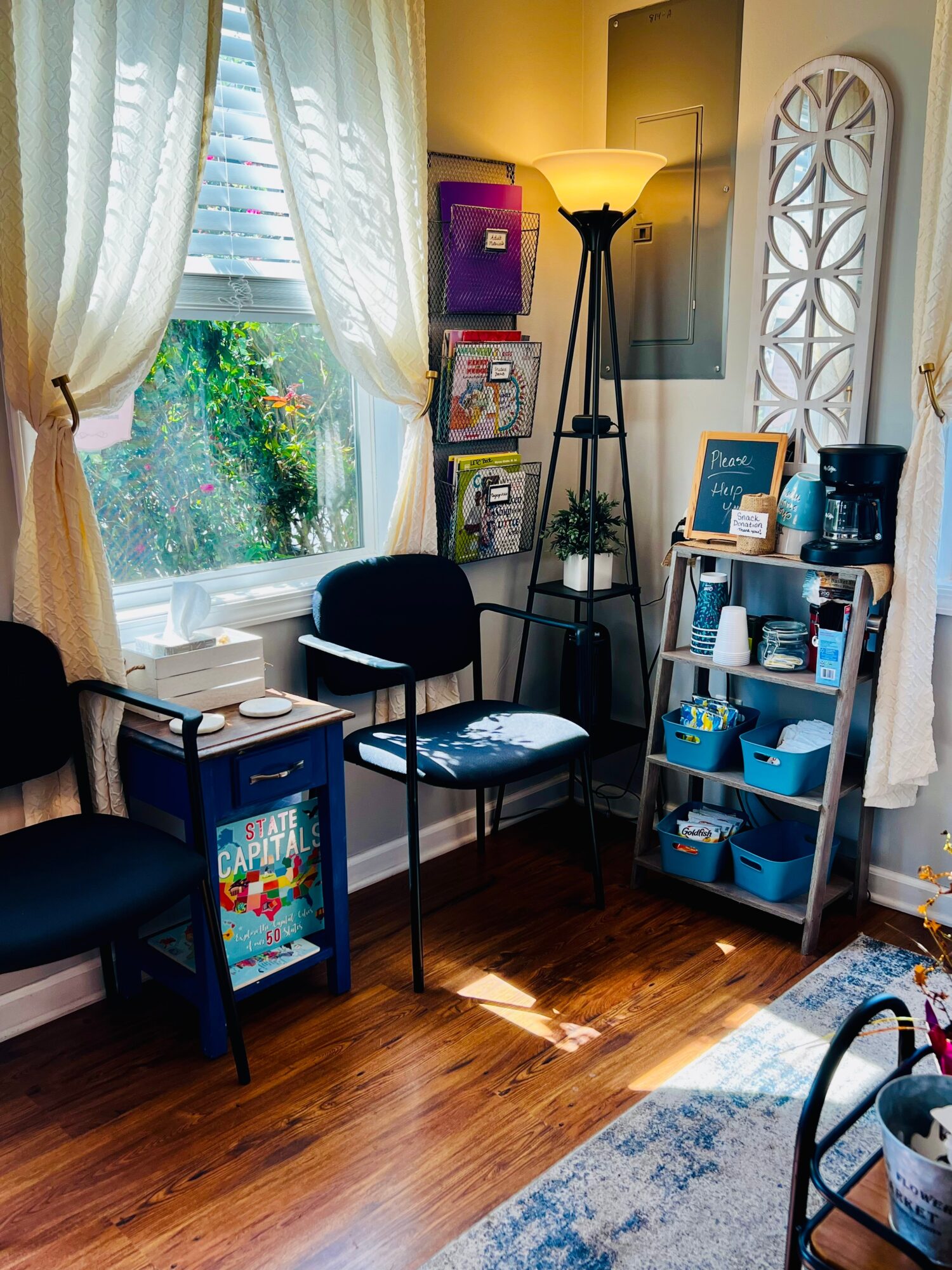 Corner of a room with two chairs, a tall lamp, a bookshelf, and a window with curtains, sunlight streaming in.