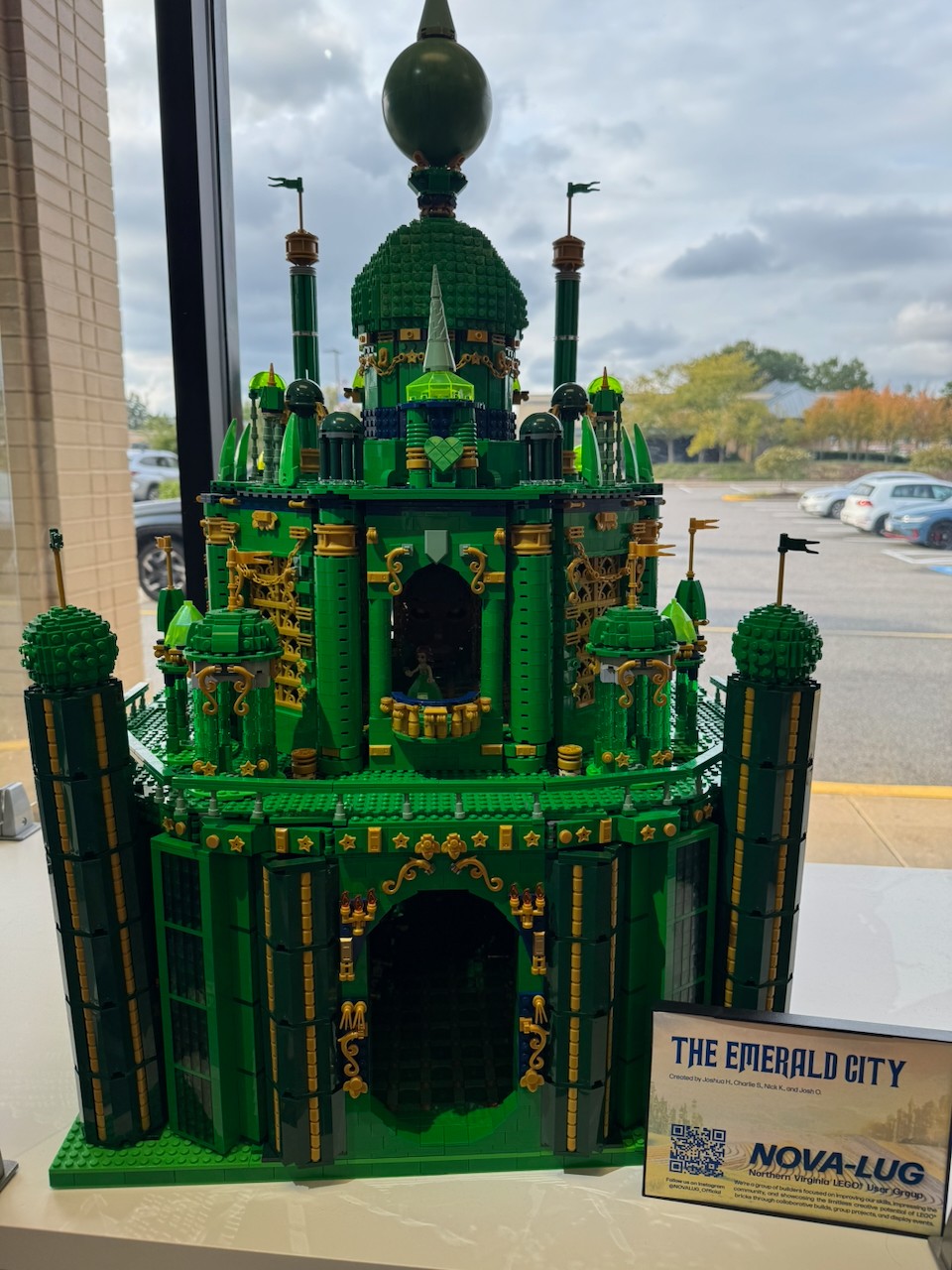 Green LEGO castle with towers, flags, and decorative elements, displayed outdoors near a parking lot and cloudy sky.