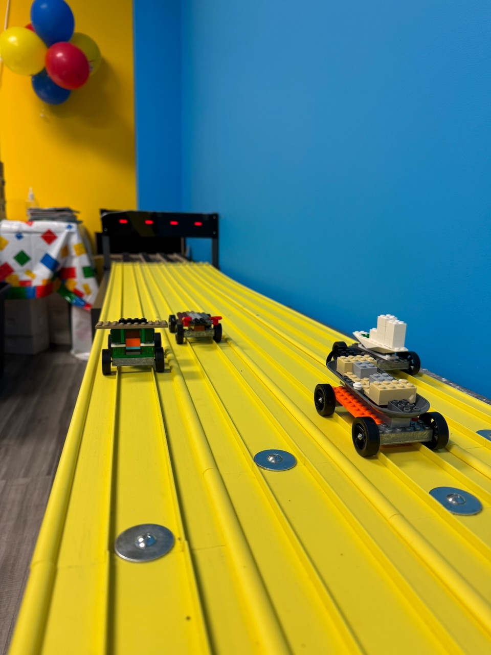 Toy cars on a yellow track with a blue wall background and balloons in the corner.