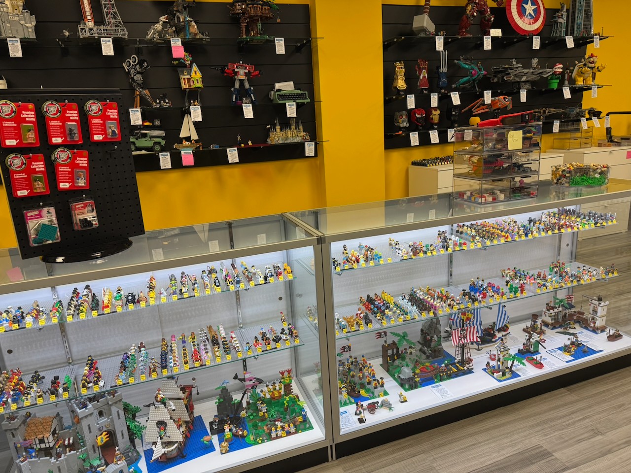 Display case filled with LEGO minifigures and sets, with shelves of collectibles behind, in a store with yellow walls.