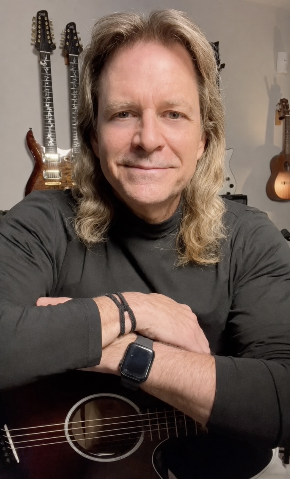 Man with long hair and a black shirt, sitting with crossed arms, in a room with guitars on the wall behind him.