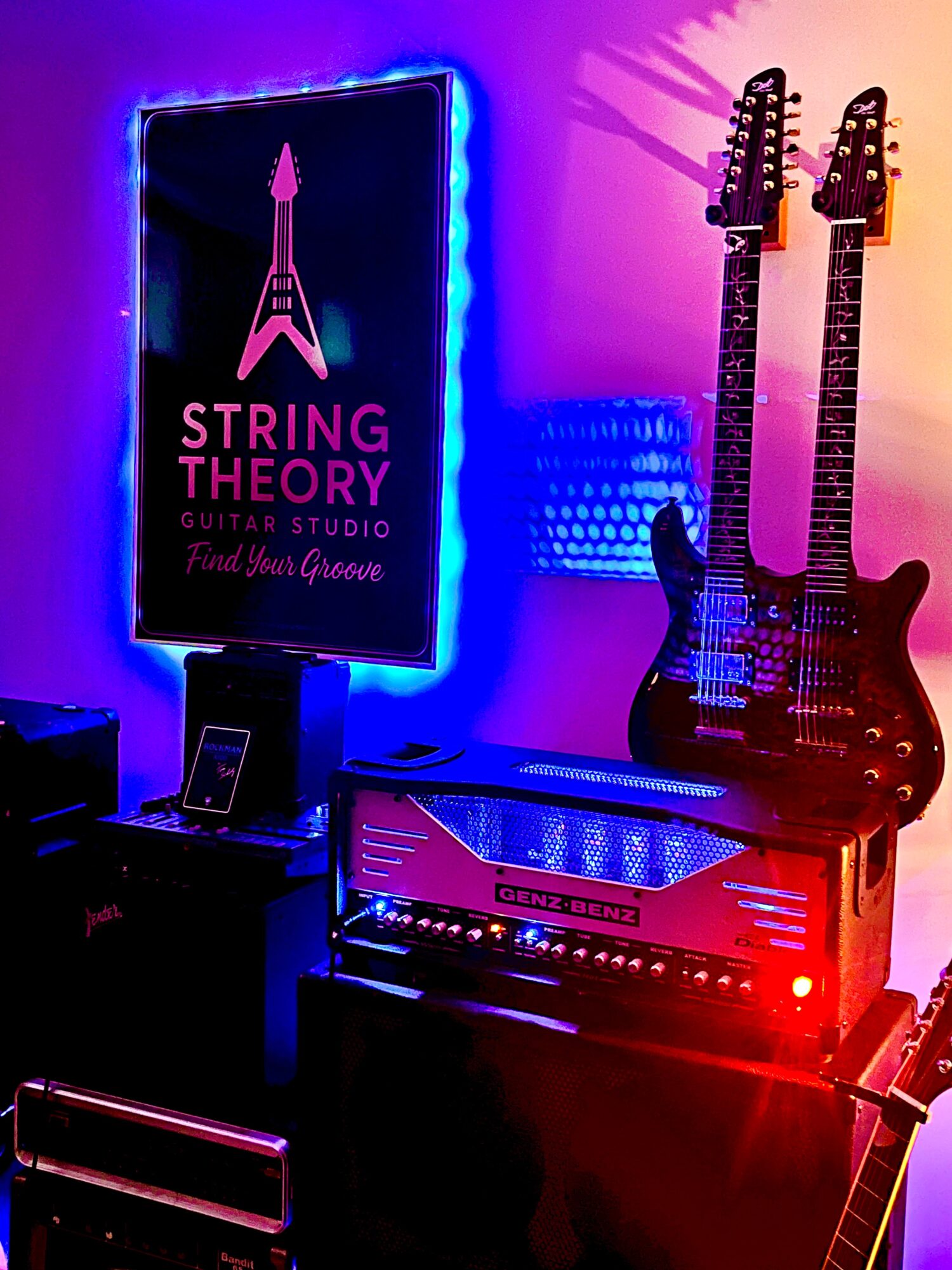 Two electric guitars on a stand, a neon sign with a guitar and text, and an amplifier with colorful lighting.