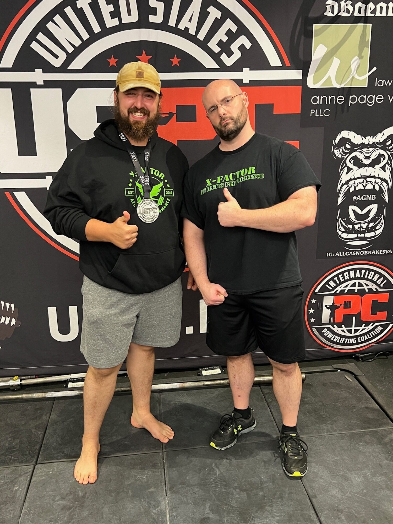 Two men standing in front of a backdrop with logos, one with a medal, both smiling and giving thumbs up.