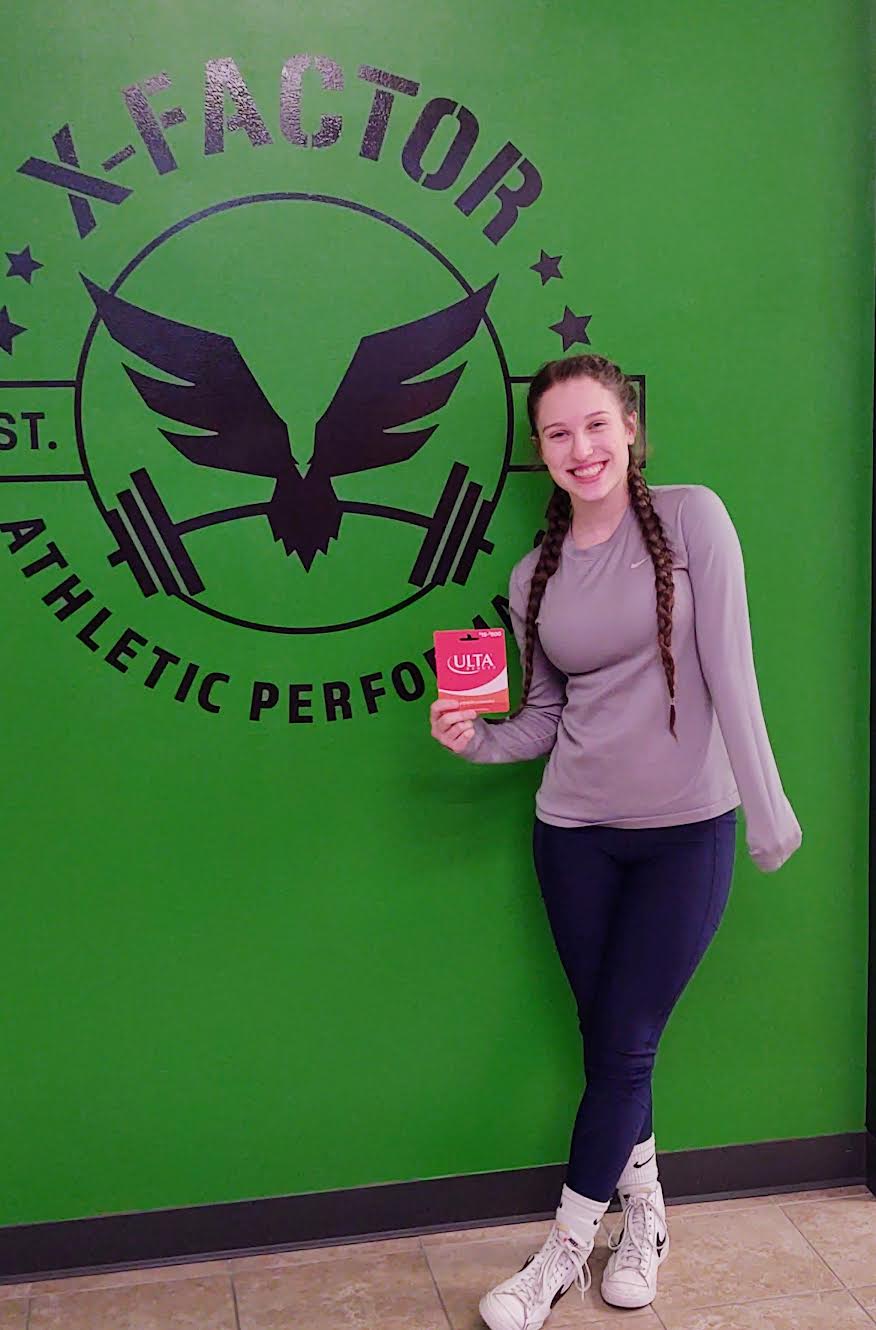 Young woman with long hair in a ponytail, smiling, holding a pink book, standing in front of a green wall with a logo.