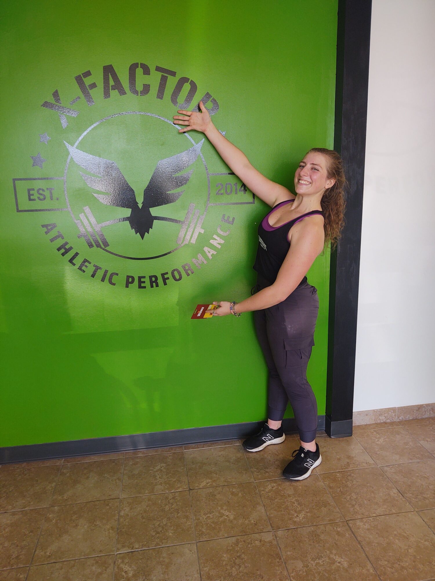 Woman in black athletic wear leaning against a green wall with a logo, holding a phone and a small object, in an indoor setting.