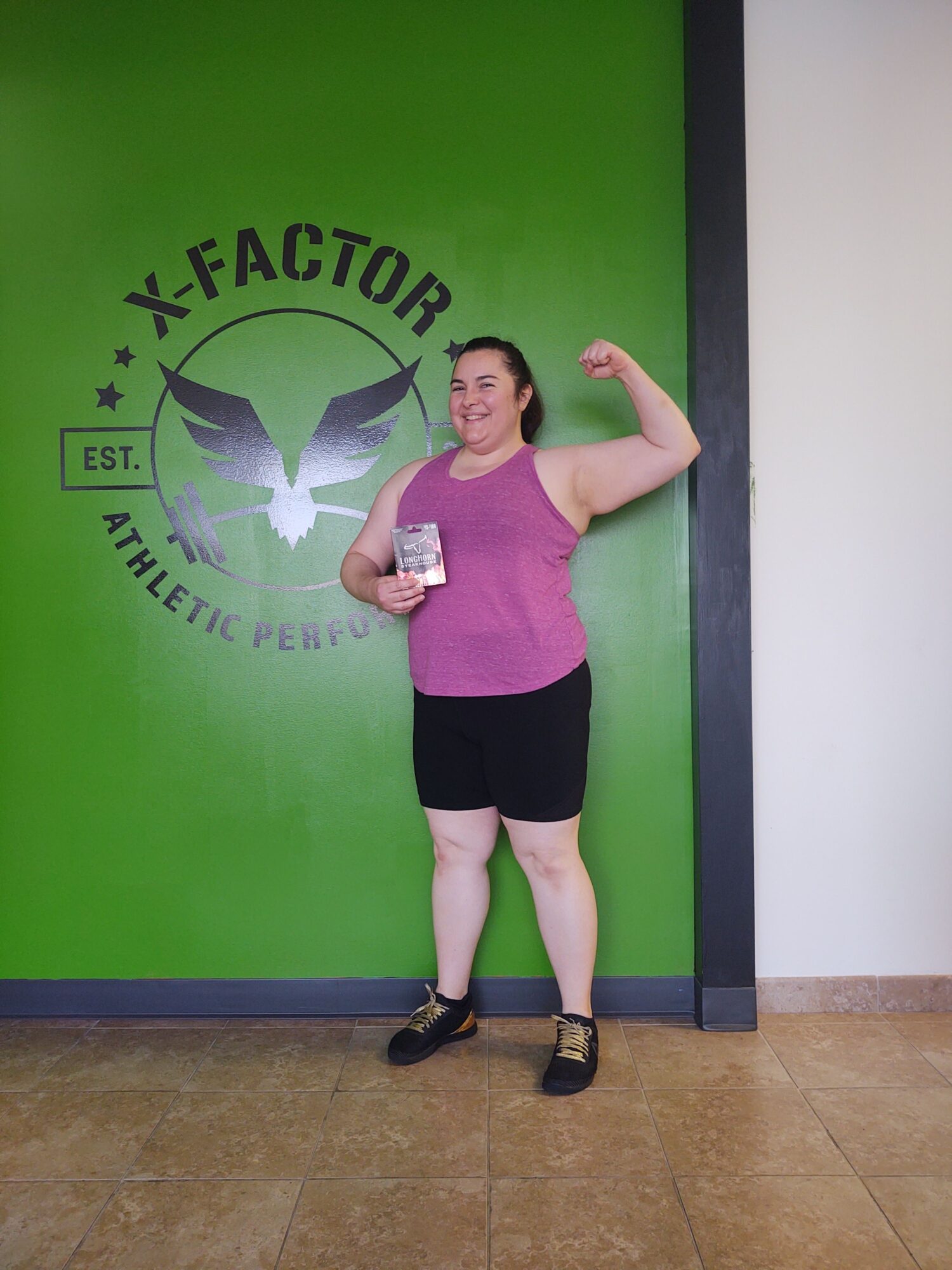 Woman standing against a green wall with a logo, holding a phone, wearing a purple top and black shorts.