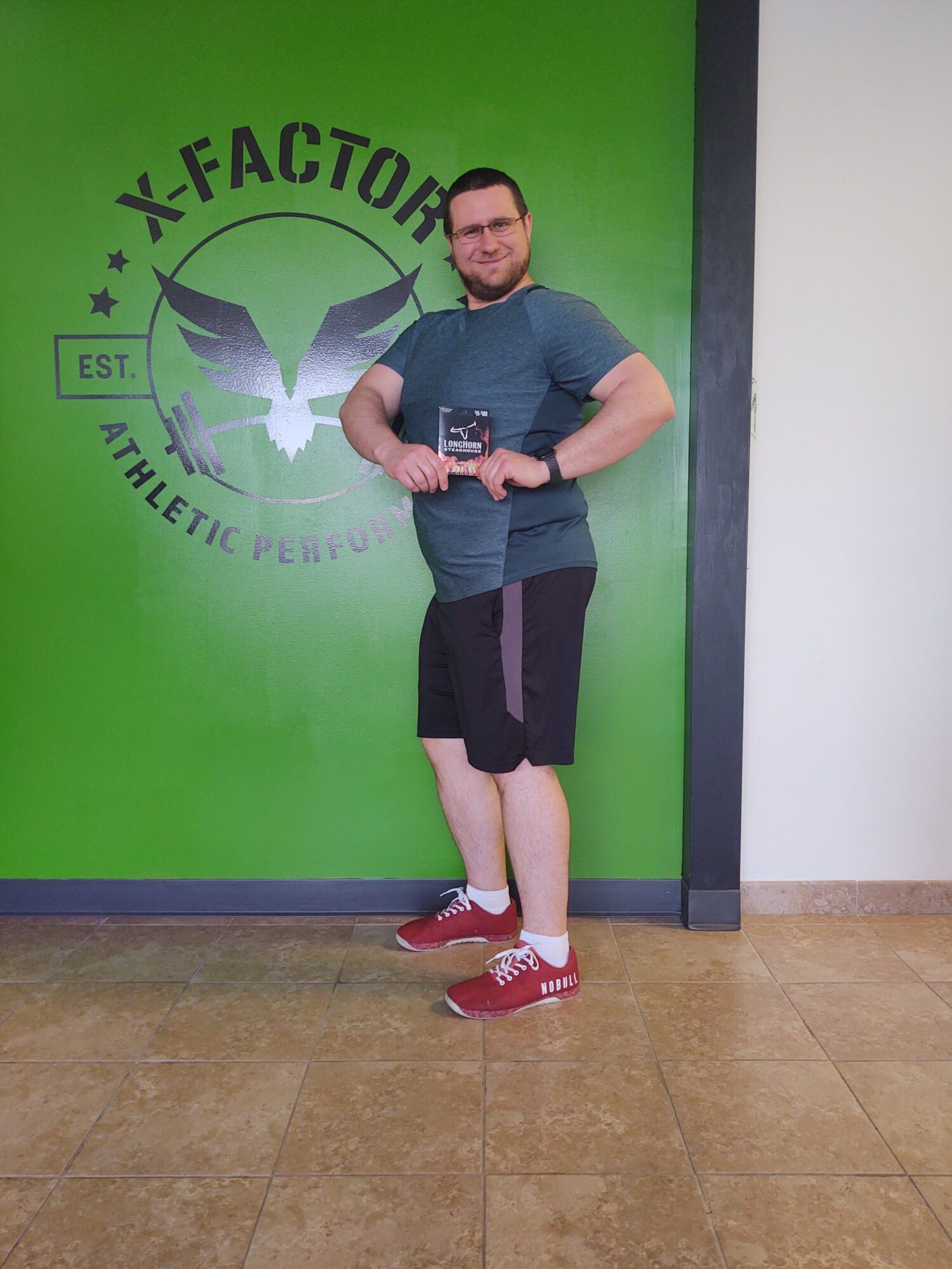 Person taking a mirror selfie in front of a green wall with a logo, wearing a gray shirt, black shorts, and red shoes.