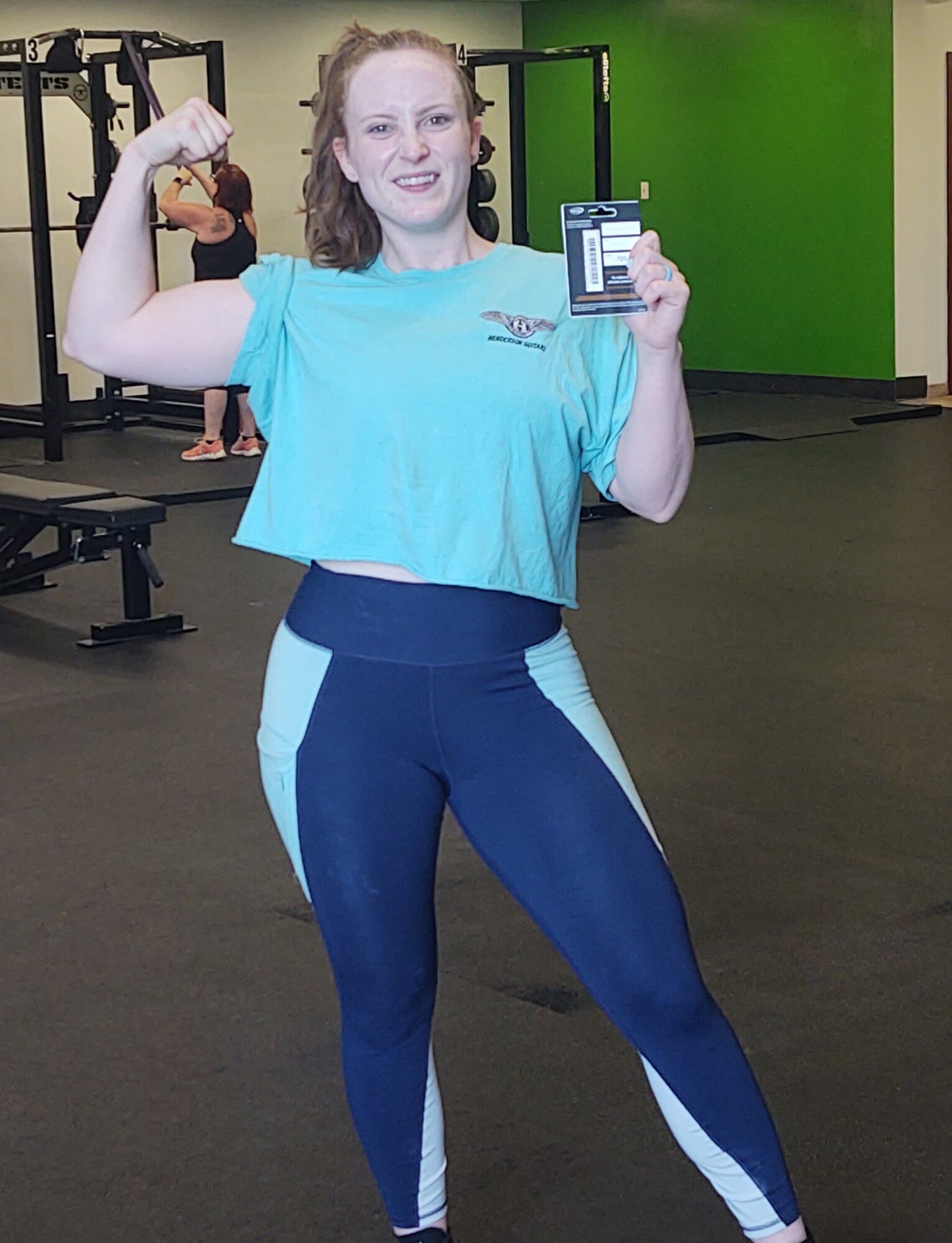 Young woman in gym holding a phone and flexing her arm, smiling, with gym equipment in background.