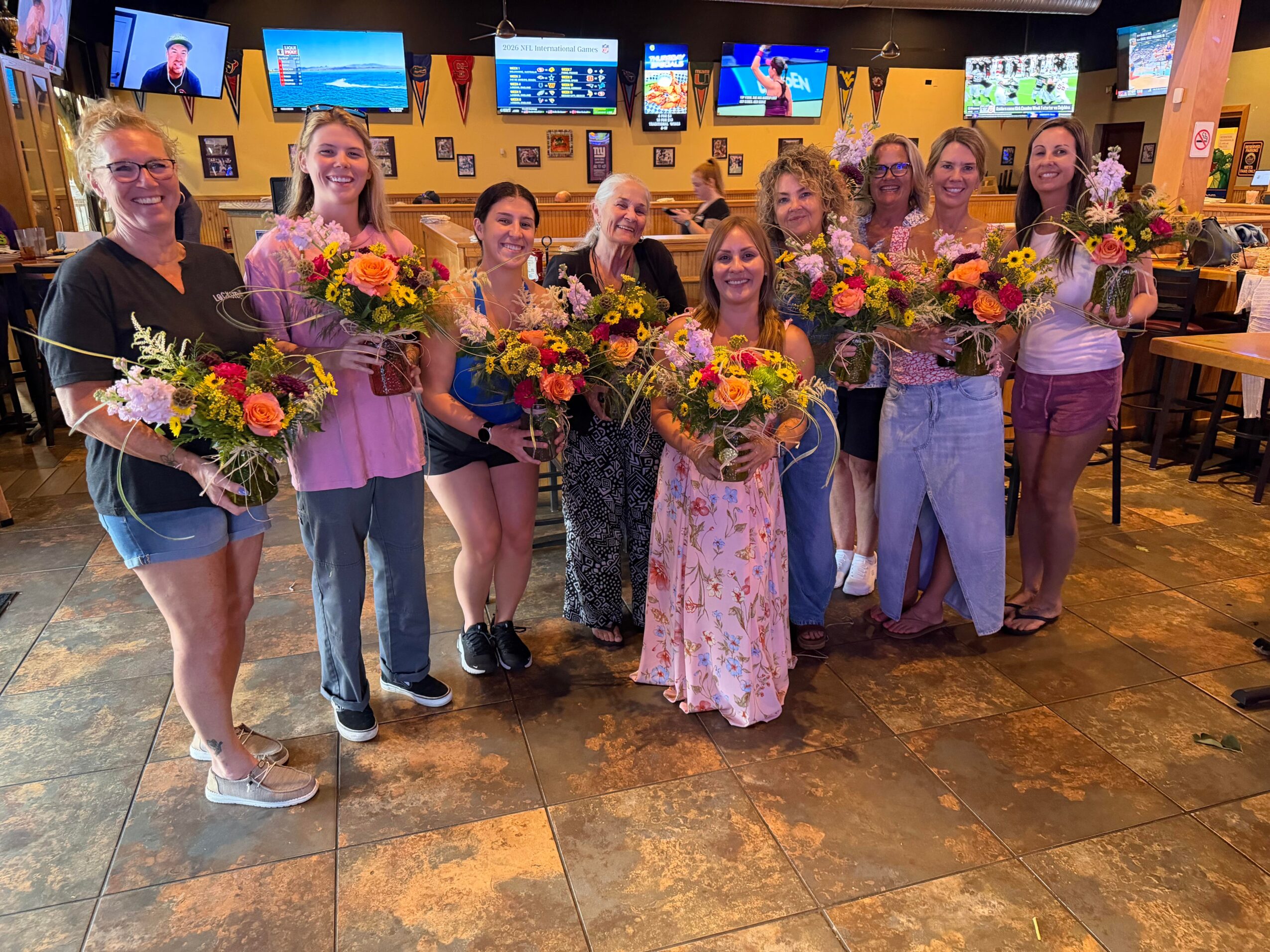 Group of ten women holding bouquets, standing inside a restaurant with multiple TV screens and dining tables.