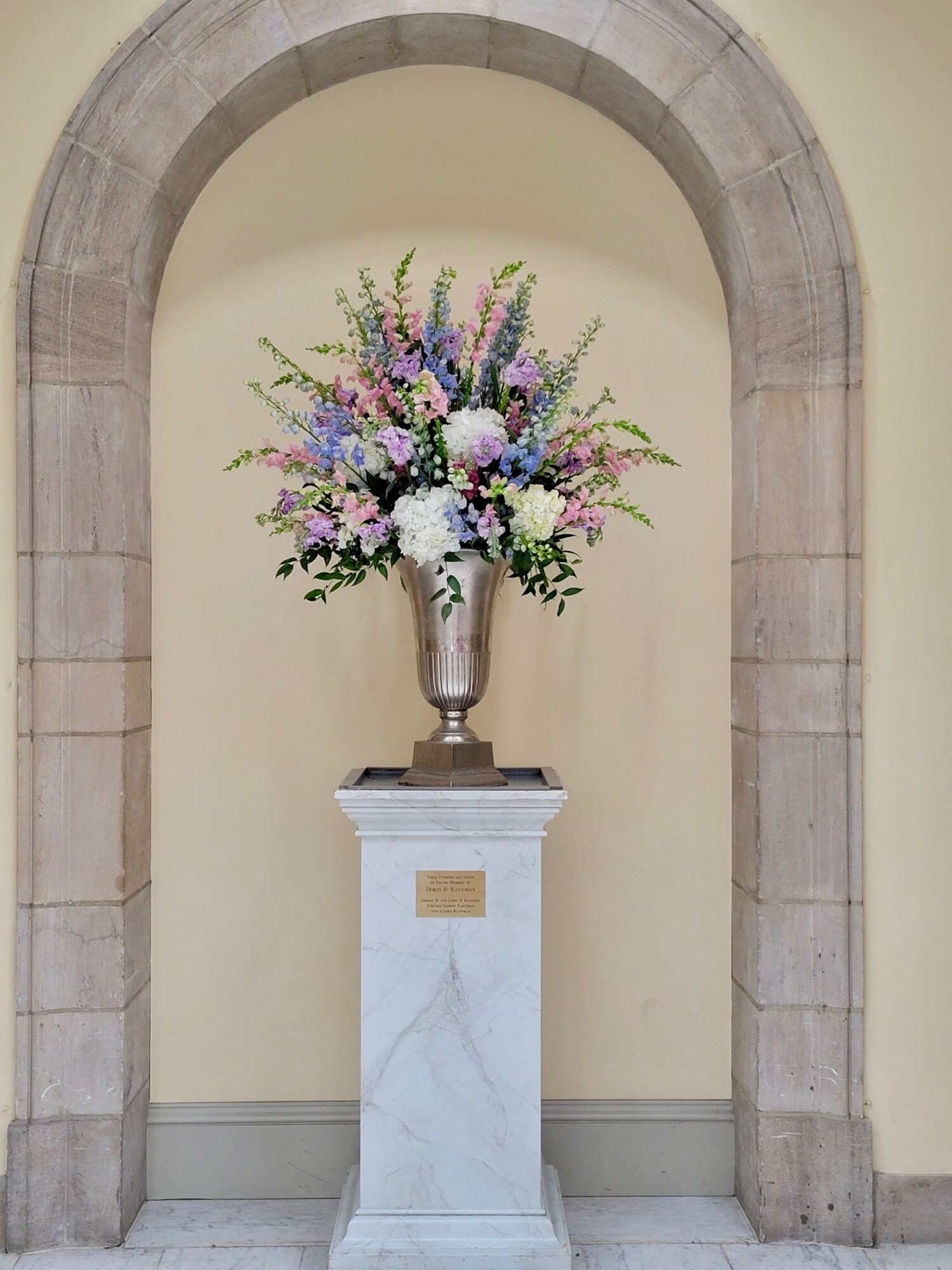 A large floral arrangement in a silver vase on a white marble pedestal inside an arched stone frame.