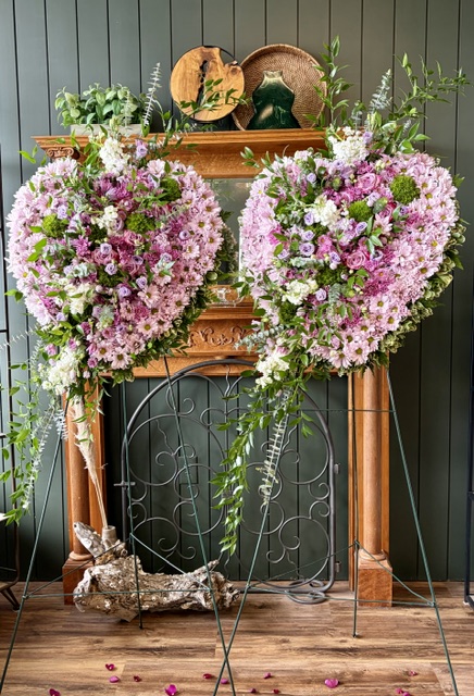 Two large heart-shaped floral arrangements with pink and white flowers on a wooden stand, with a decorative metal frame below, against a dark wall.