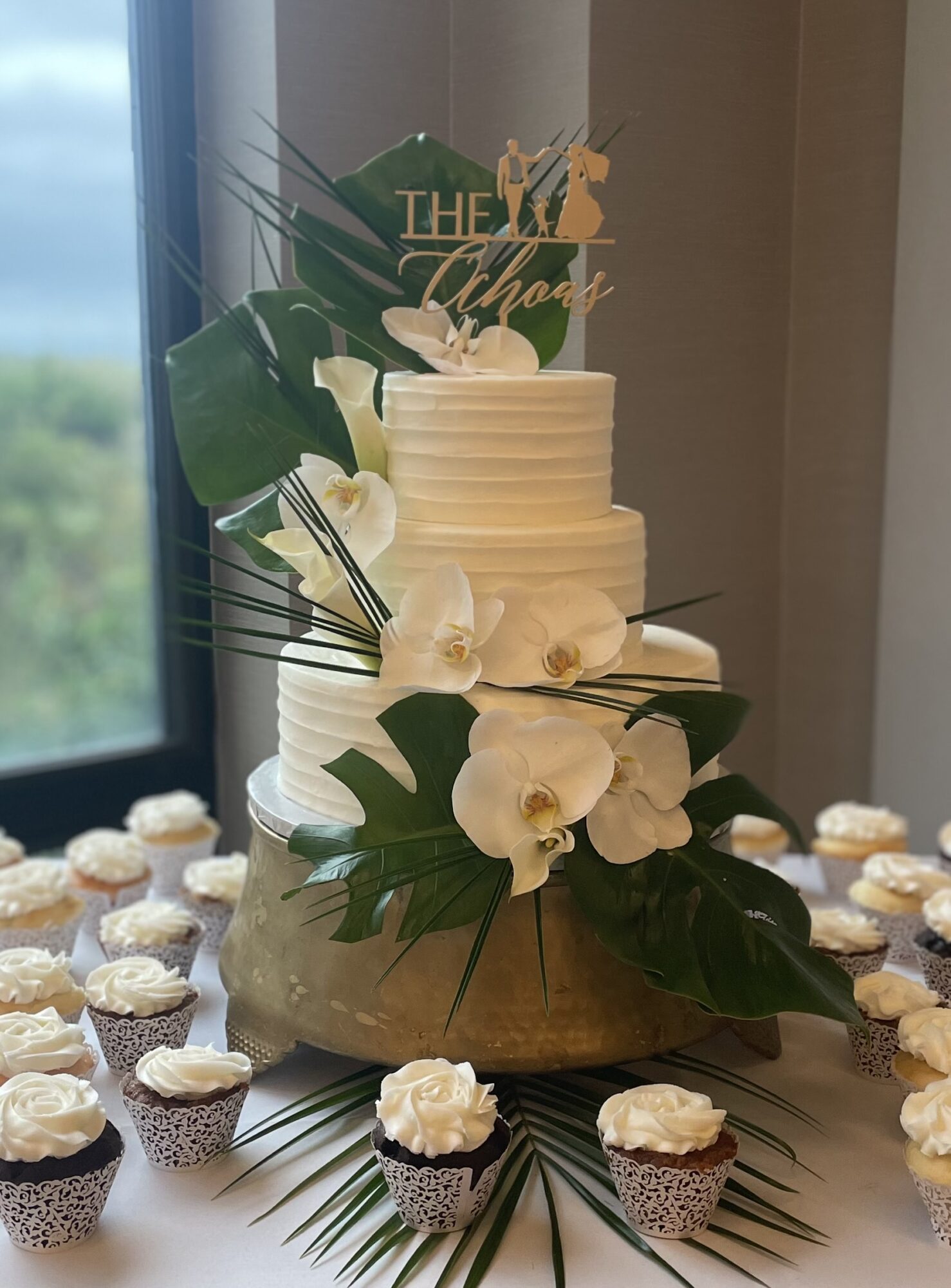 A white tiered wedding cake decorated with white flowers and green leaves, surrounded by cupcakes on a table.