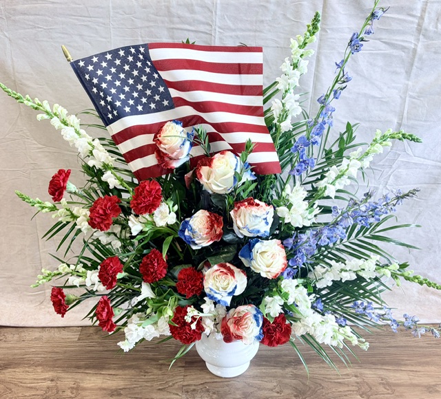 Flower arrangement with red, white, and blue flowers, an American flag, and green foliage in a white vase on a wooden surface.