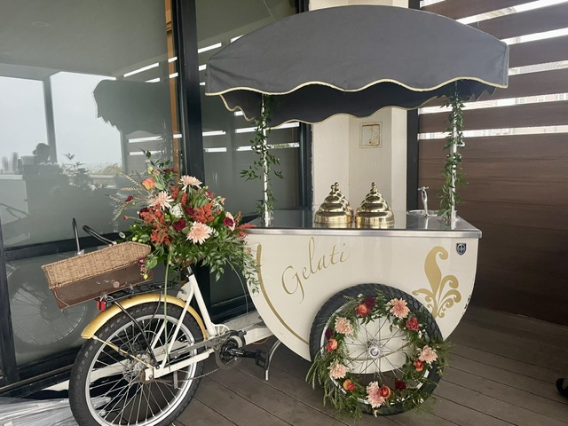 Gelato cart with umbrella, decorated with flowers and a bicycle, positioned outdoors near a glass window.