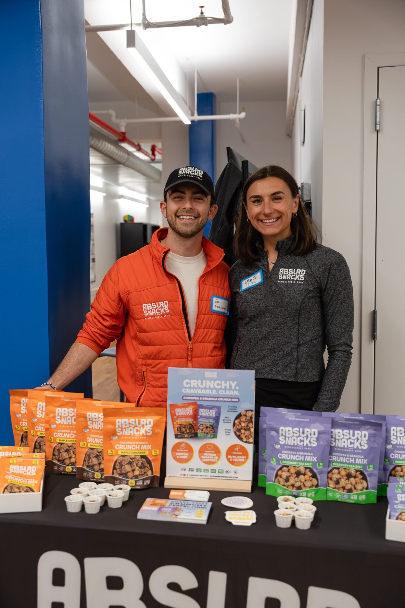 Two smiling people stand behind a table with snacks and products, in an indoor setting with blue and white walls.