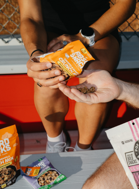 Person holding a snack bag, another hand reaching for a cookie, table with snack packages, outdoor setting.