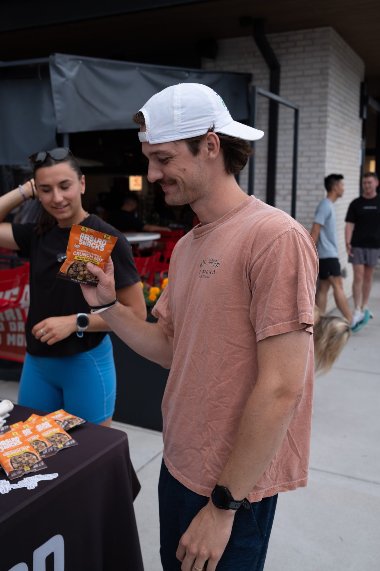 Young man in a white cap and pink T-shirt smiling while holding a snack package outdoors, with people in the background.