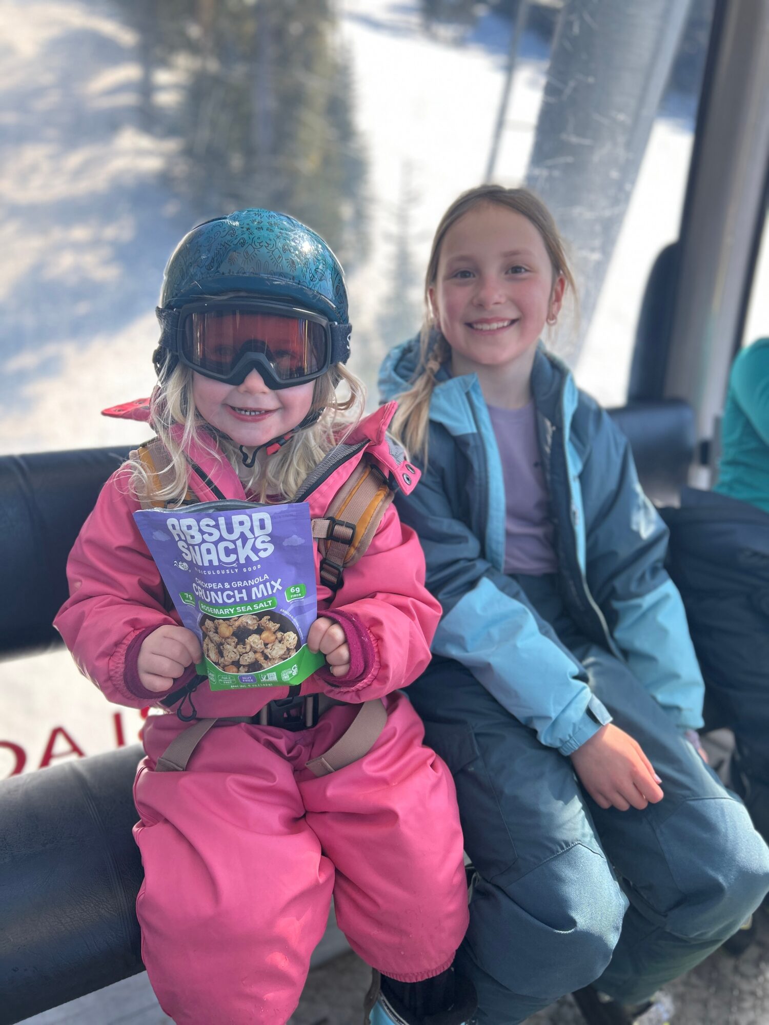 Two children sitting on a ski lift, smiling, with snow and trees in the background.