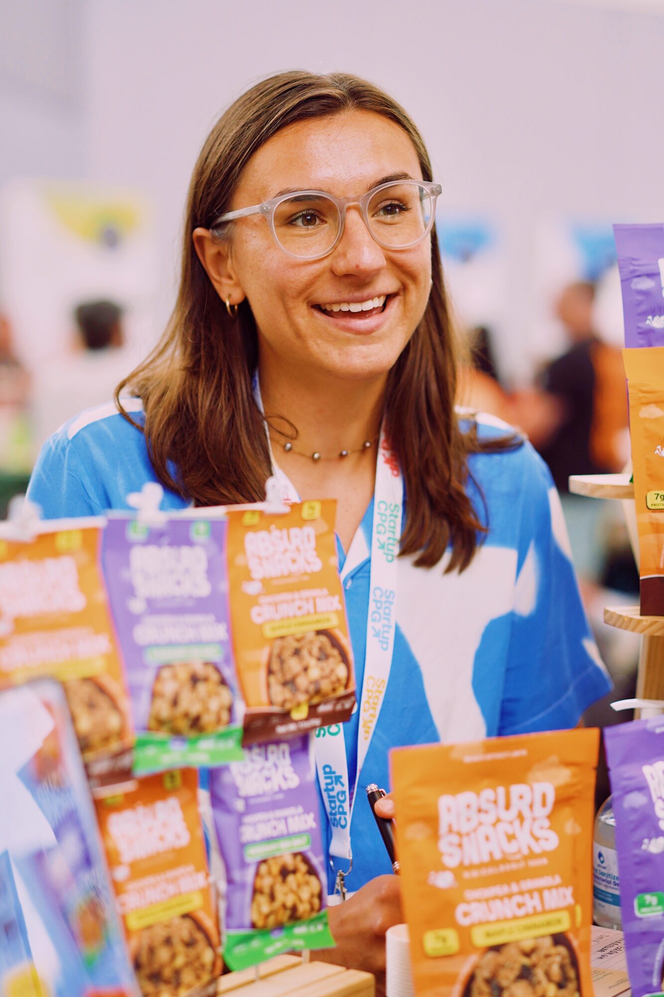 Woman with glasses smiling at a booth with snack packages, wearing a blue and white patterned shirt, in an indoor setting.