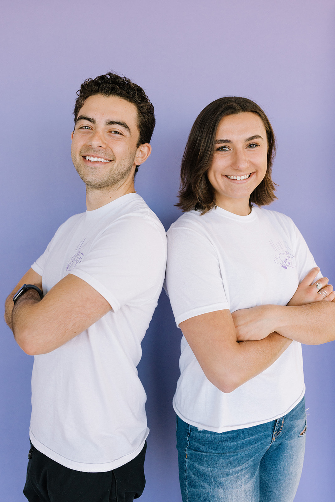 Two smiling people stand with arms crossed against a purple background.
