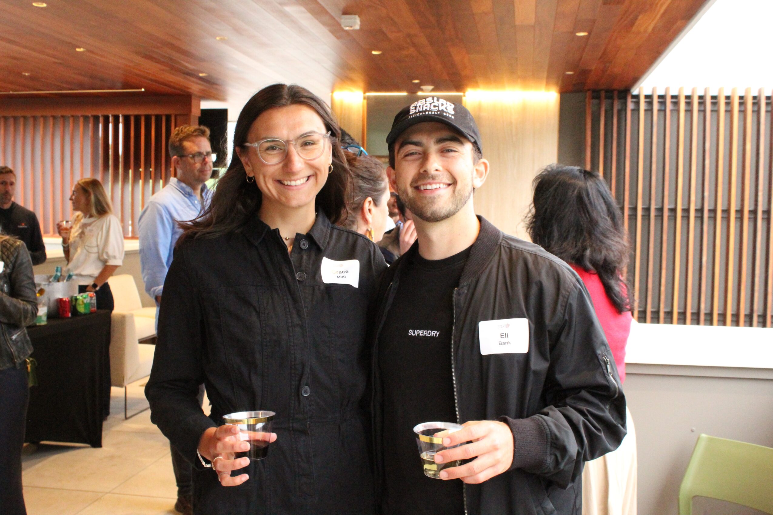 Two smiling people holding drinks at an indoor social event, with other guests in the background.