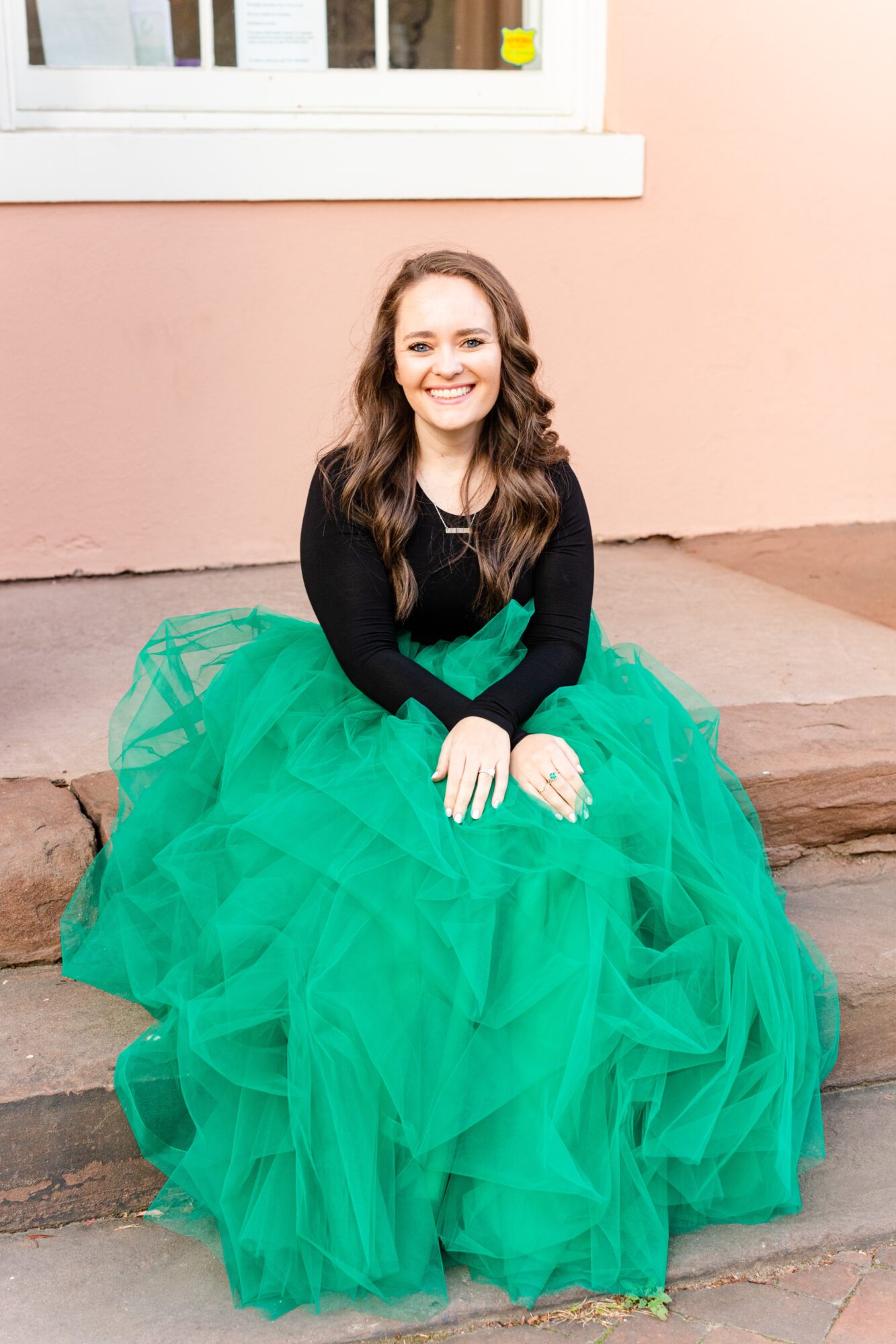 Young woman with long brown hair smiling, sitting on steps in a black top and green tulle skirt, against a pink wall.