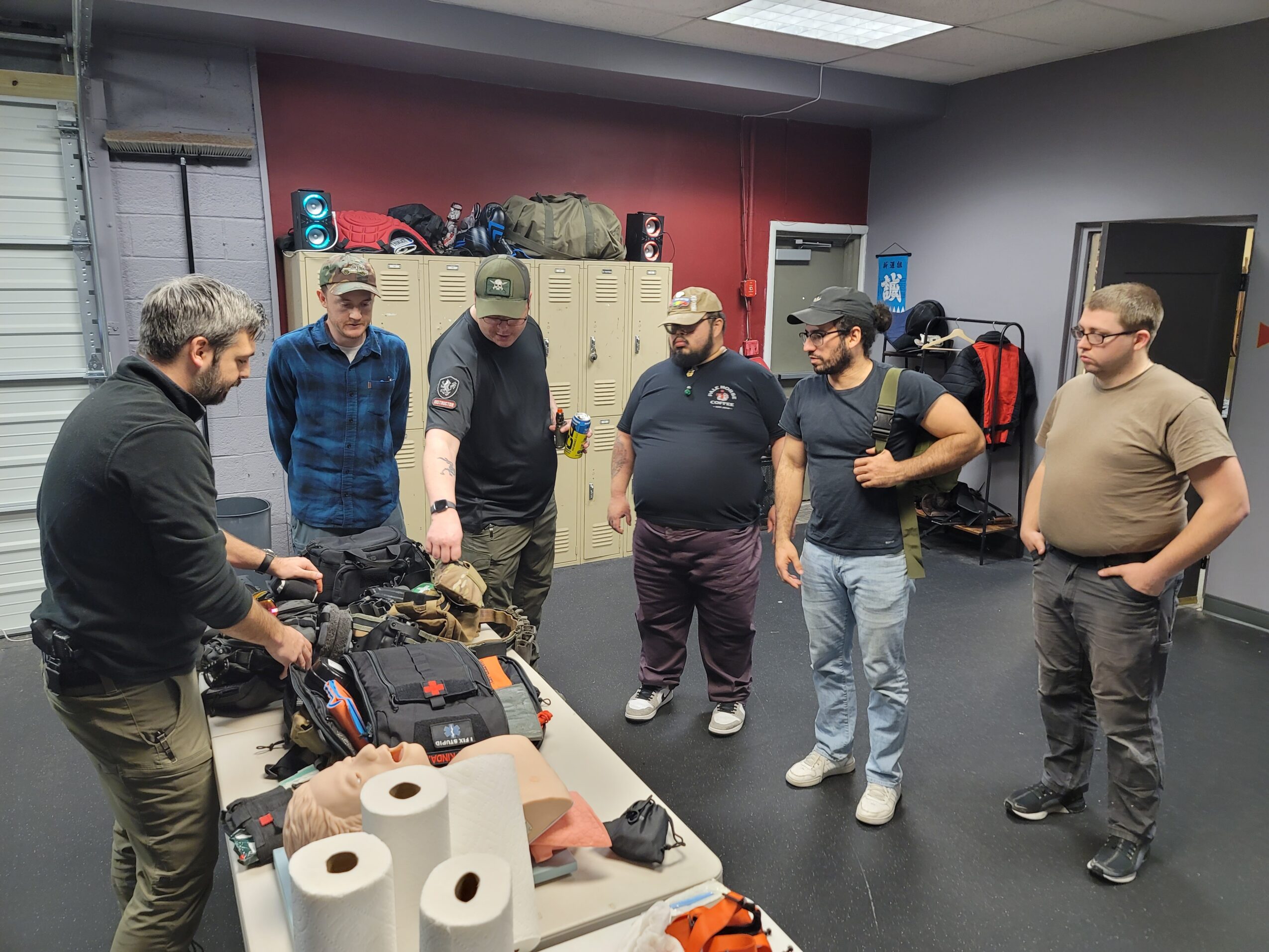 Six men stand around a table with tactical gear and equipment in a room with lockers and backpacks.