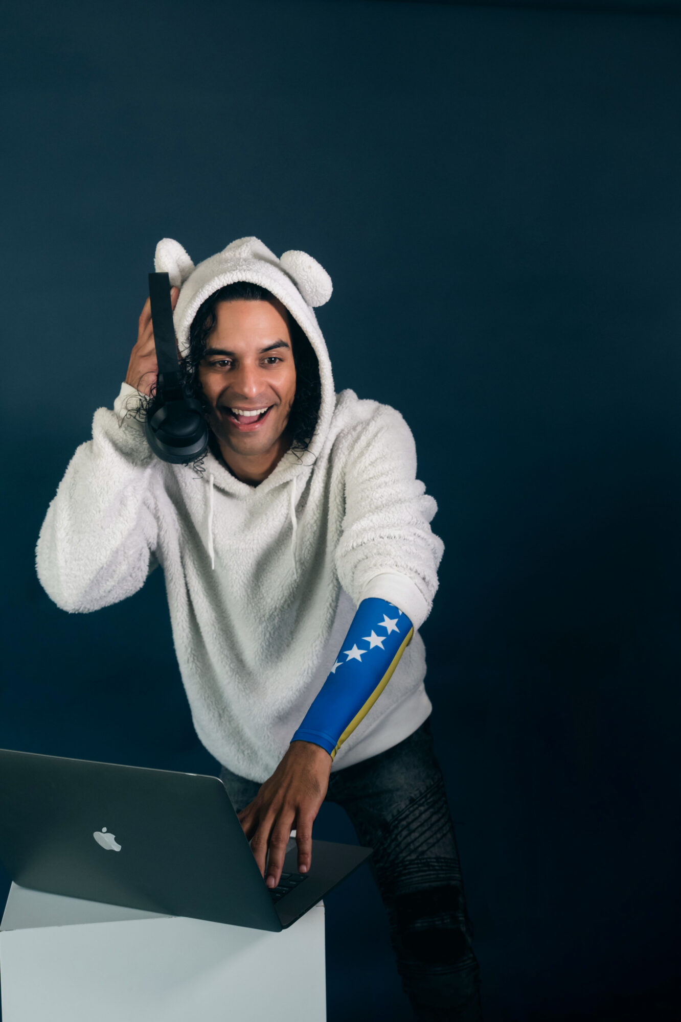 Person smiling wearing a bear hat and a white hoodie, sitting at a desk with a laptop, dark background.